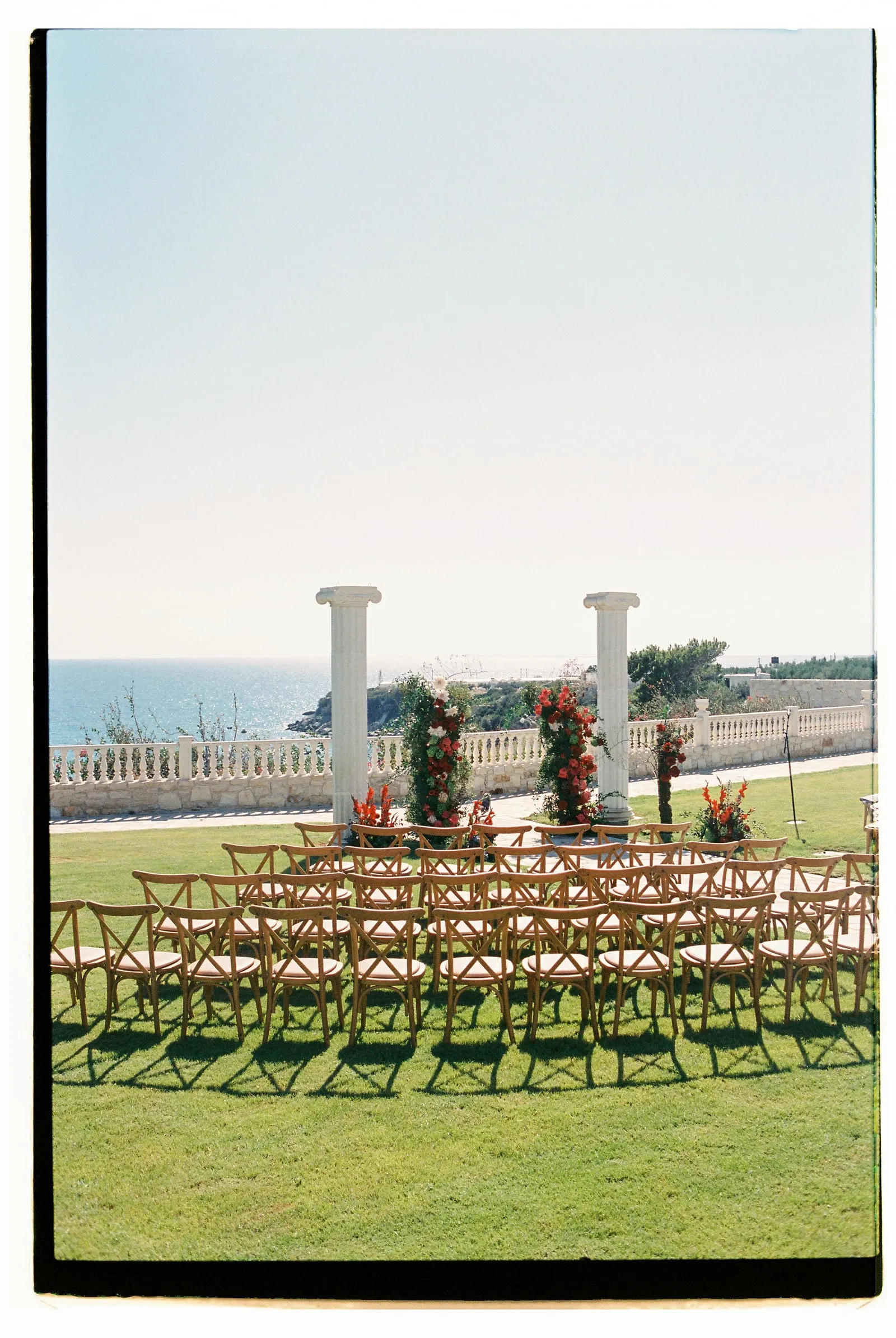Rows of wooden cross-back chairs with cushioned seats fan out across a lawn toward a ceremony spot framed by two tall white stone columns. Floral installations of red and white blooms, likely garden roses and gladioli with green foliage, stand beside the columns, with a pale balustrade and the blue Aegean Sea beyond. This wedding ceremony set-up at Wine Dark Sea Villas, on Crete in Greece, is bathed in soft, bright sunlight under a clear sky.