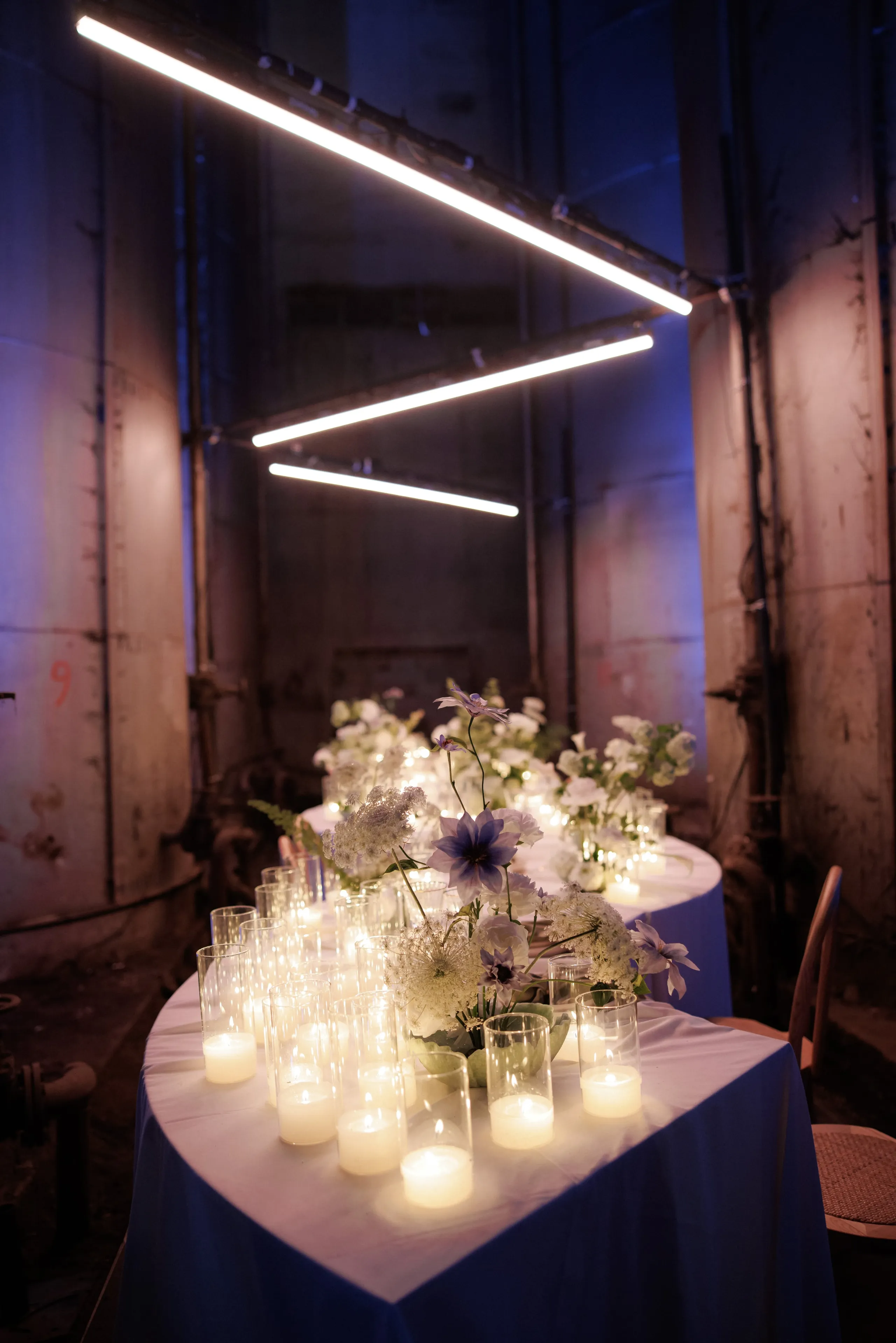 A long curved wedding banquet table in a industrial storage hall in Rethimno, Crete, Greece, draped in a crisp white cloth, with clusters of white queen anne's lace, white stock and deep violet-blue anemones or clematis-like blooms running down its centre. Rows of glass votive holders with lit candles glow in front, while overhead, angled strip lights and the cool blue wash on the concrete walls create a moody, modern atmosphere.