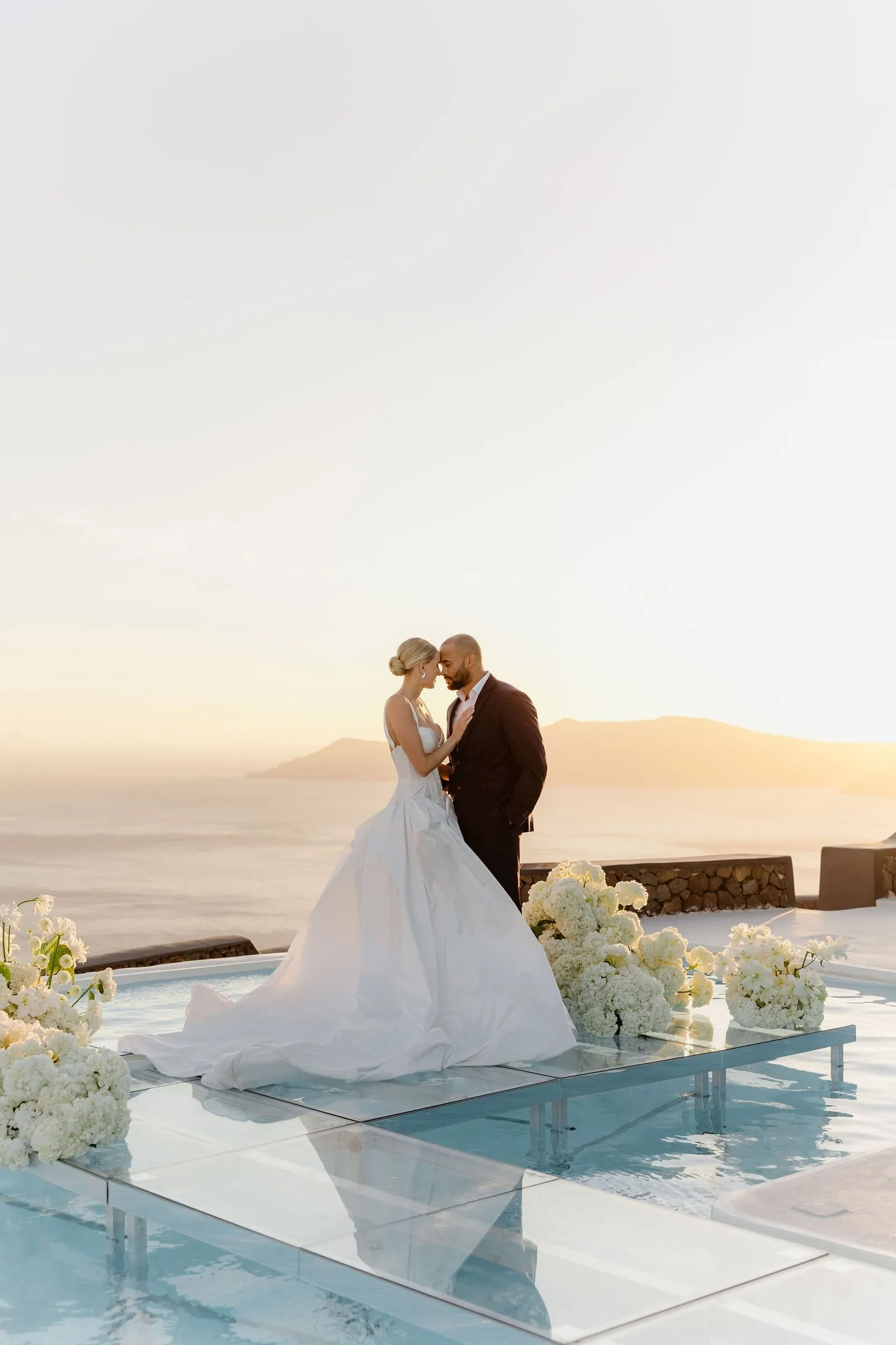 A bride in a flowing white ballgown with a long train and her hair in a soft low bun stands close to a groom in a dark suit, foreheads almost touching, on a glass walkway above an infinity pool at Aenaon Villas in Santorini, Greece. Clusters of white hydrangeas sit along the edges of the glass path, and the pool water reflects the pale sky. Behind them the sea fades into hazy hills in warm golden sunset light, making this a wedding moment of quiet intimacy.