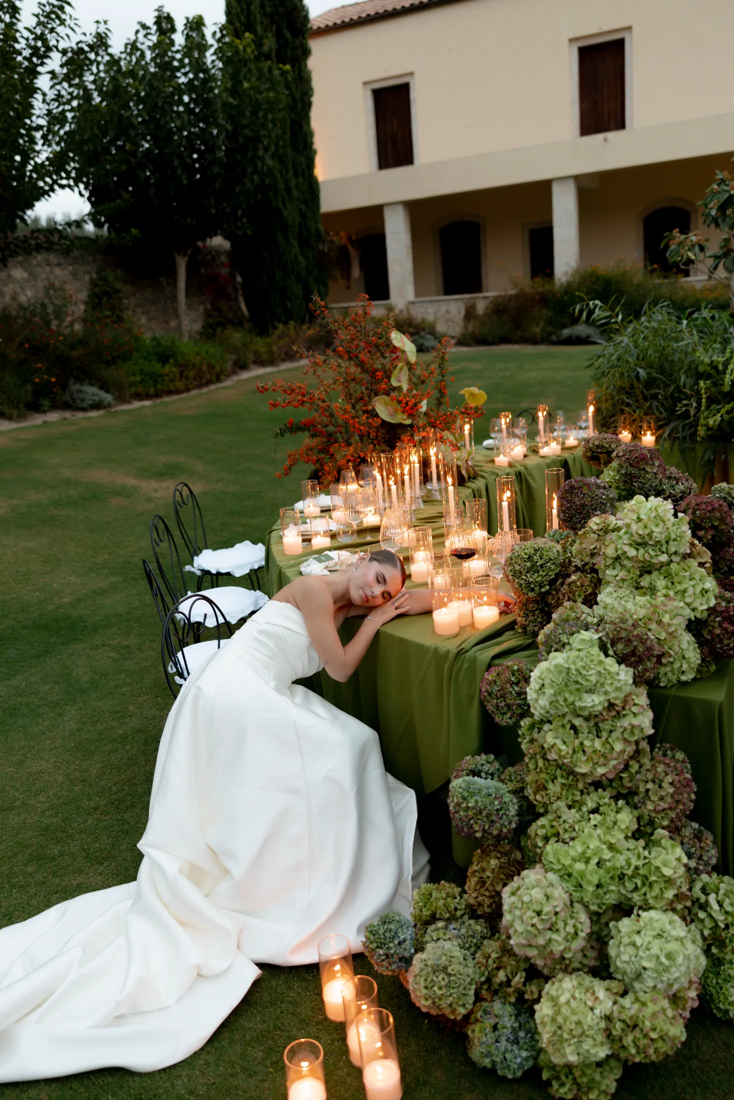 A bride in a white strapless gown with a long train rests her head on her arm at a long table draped in olive-green linen on the lawn of Ktima Roxani estate in Armeni, Crete, Greece. Pale green and mauve hydrangeas heap up around the table's end, while a tall arrangement of orange-red berried branches and broad leaves stands at the far end. Clusters of lit candles in glass holders line the table and the grass, and a stone villa with arched windows glows in the soft evening light.