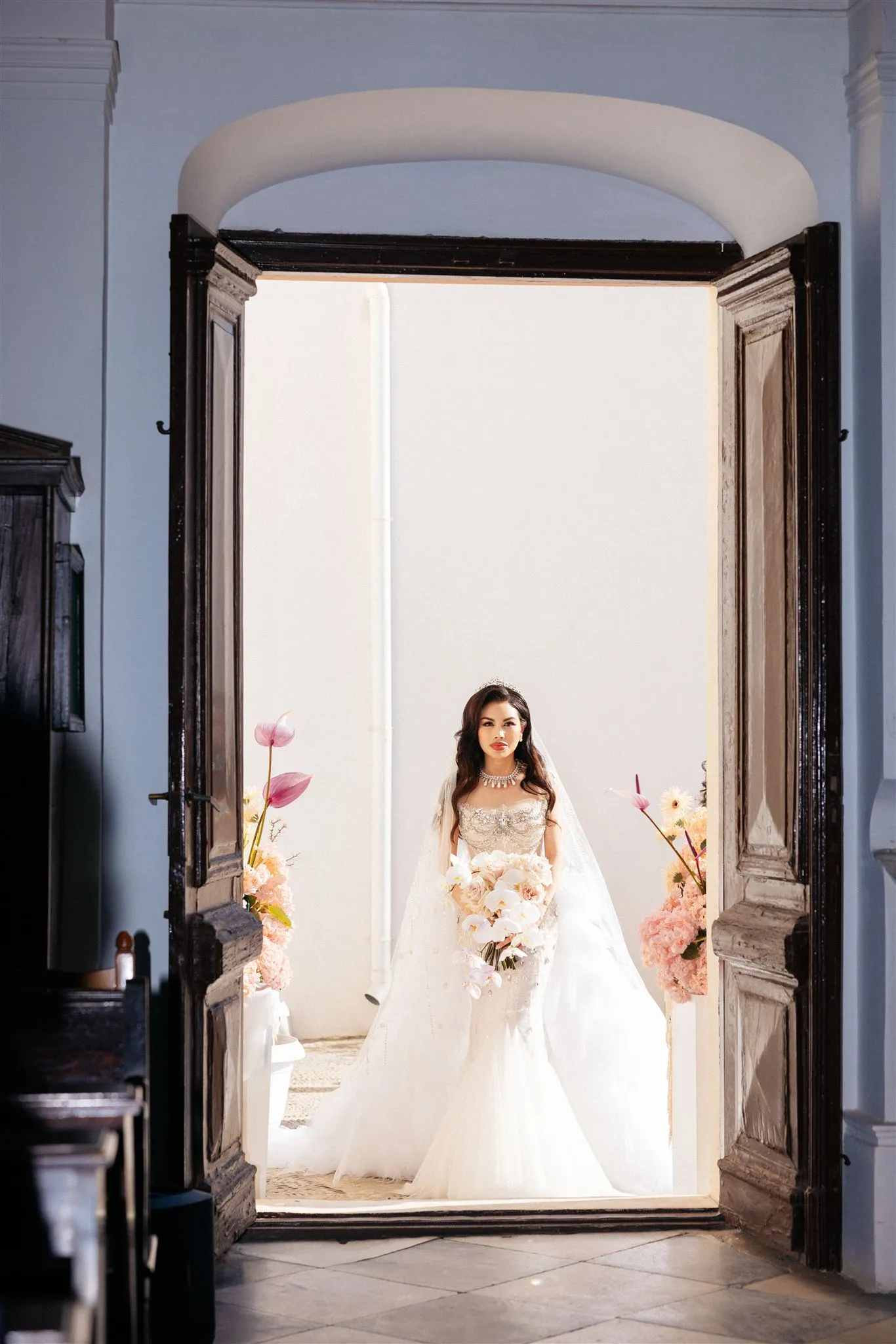 A bride stands framed in the open doorway of a pale blue church at Islet in Santorini, Greece, bathed in bright light from the courtyard beyond. She wears a fitted, beaded white gown with a long veil and a jewelled necklace and tiara, holding a cascading bouquet of white orchids and blush roses. On either side of the wooden doors, pink anthurium and peach hydrangea arrangements frame her, and the dark church pews sit in soft shadow in the foreground.