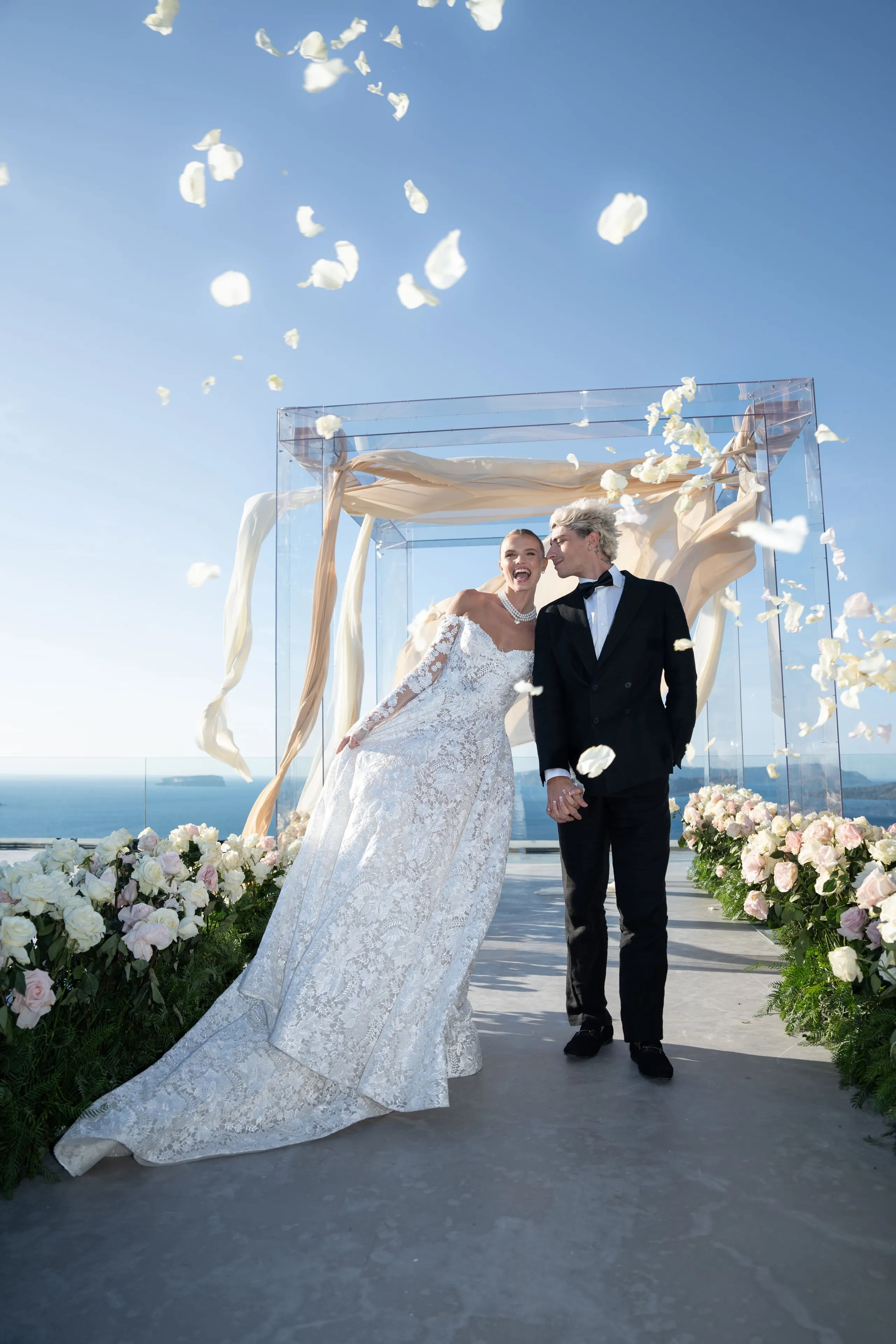 A bride in a lace gown with long sleeves laughs joyfully beside her groom in a black tuxedo and bow tie, as white rose petals drift down around them. Behind them stands a clear acrylic arch draped with champagne-coloured chiffon, with the blue Aegean sea stretching under a cloudless sky. Lush beds of white and blush roses line the path, a wedding moment on the terrace at Cavo Ventus Luxury Villa in Santorini, Greece.