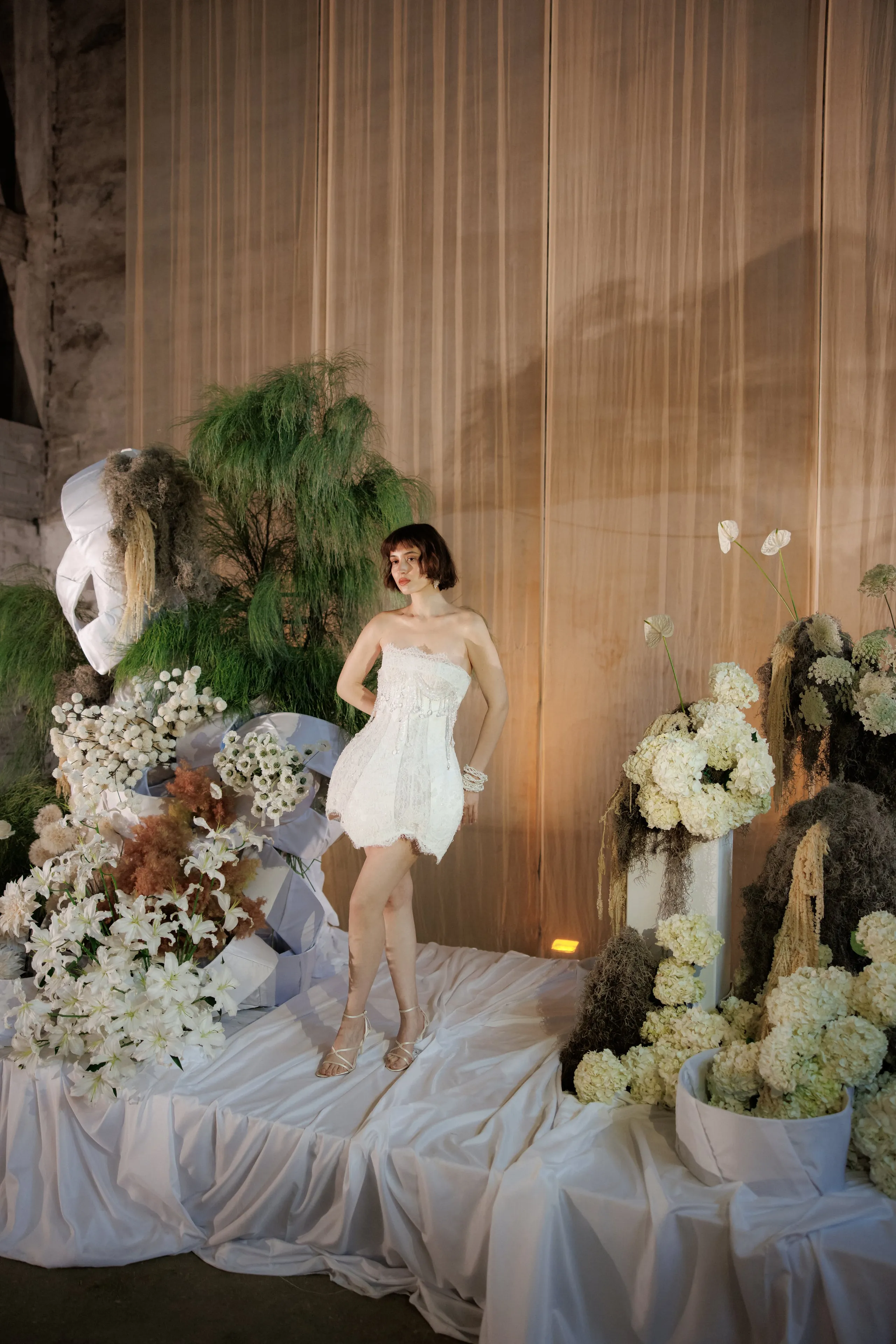 A bride in a short, strapless white lace dress stands on a draped white satin platform, framed by a lush wedding floral installation at Rethimno, Crete, Greece. Clusters of white lilies and cream hydrangeas spill from large white containers, while feathery green asparagus fern and dried wheat-toned plumes rise behind her against a pale, softly lit curtain.