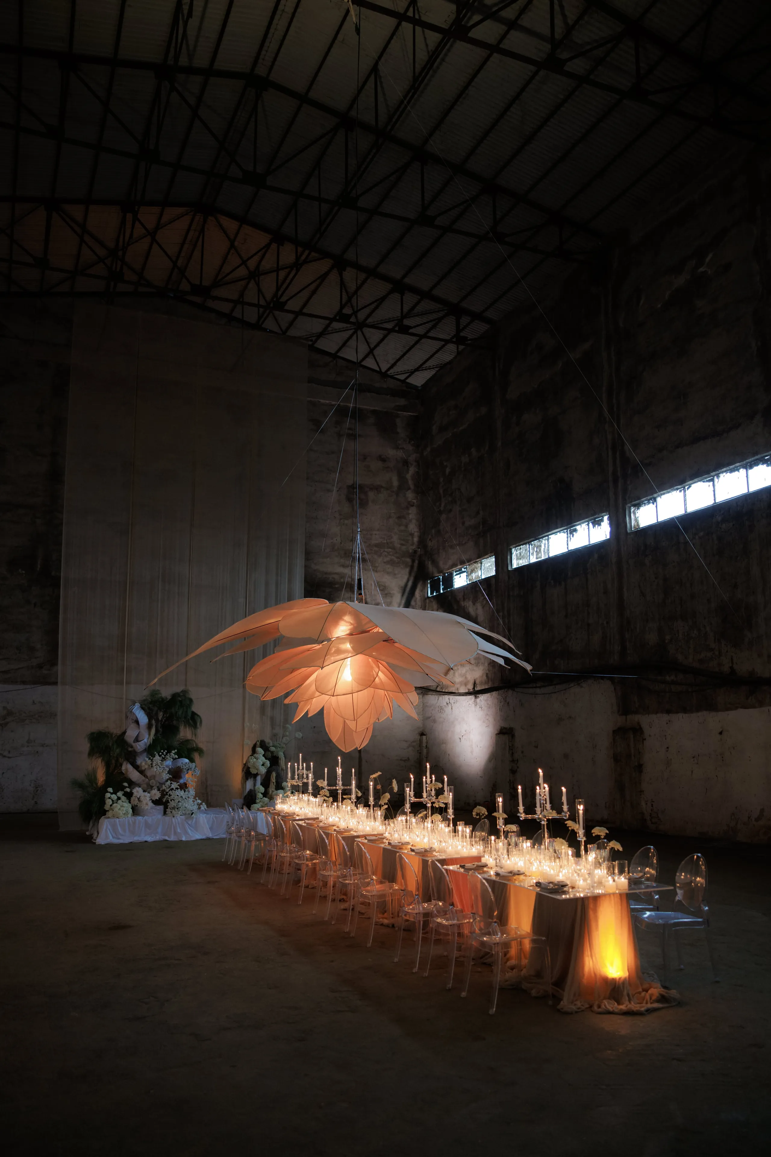 A long wedding table set for dinner inside a vast, dim industrial hall in Rethimno, Crete, Greece, with clear acrylic chairs lining both sides. Beneath a huge pale, petal-shaped lamp hanging from the rafters, rows of tall candelabra and glowing candles run down the table over amber and sand-coloured linen, and a warm glow spills from beneath the cloth. At the far end, white hydrangea-style blooms and dark green foliage gather around a sculptural pale form beside a flowing fabric backdrop.