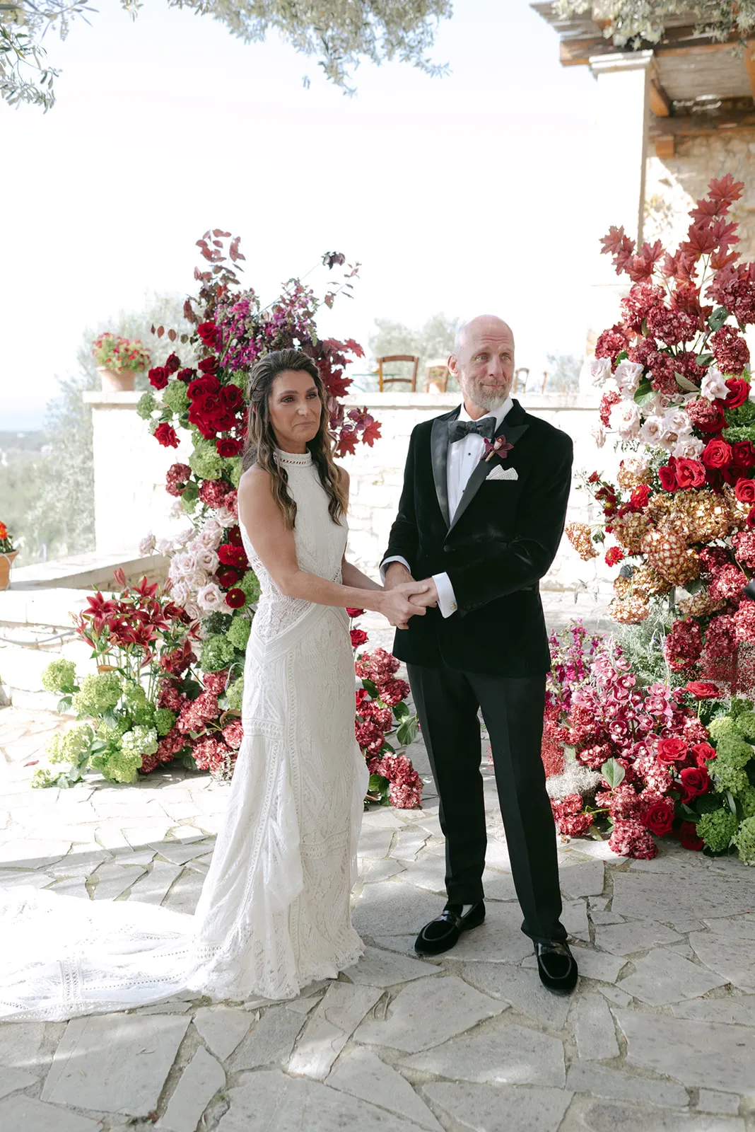 A bride in a lace high-neck gown and a groom in a black velvet tuxedo hold hands before a wedding arch of red garden roses, cream roses, green hydrangeas and deep burgundy foliage, with tall pink and red spires rising behind them. This wedding flower installation at Agreco Farm in Greece sits on a pale stone terrace with olive trees and hills beyond.