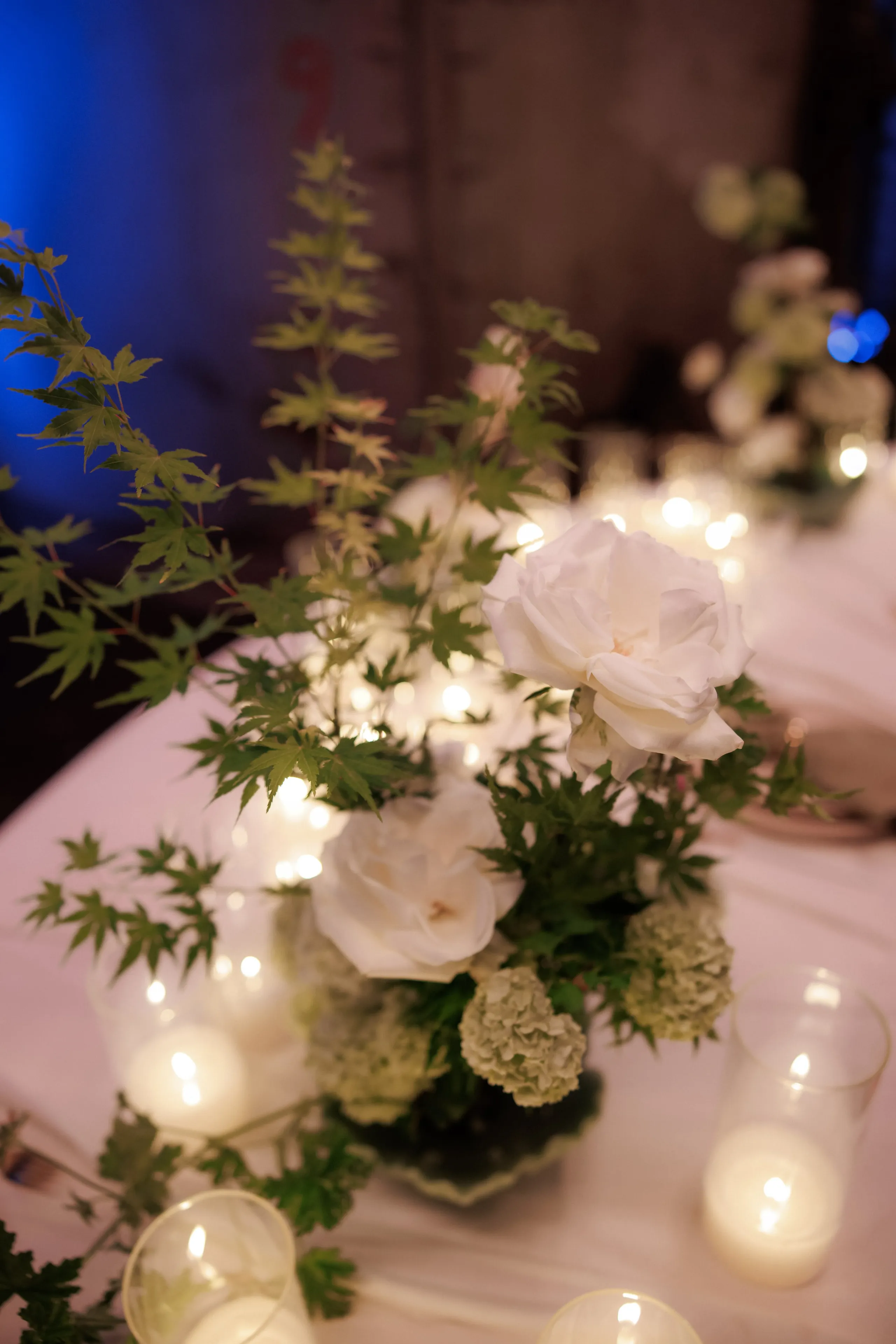 A low wedding table centrepiece in Rethimno, Crete, Greece: two creamy white garden roses rise above feathery green Japanese maple sprigs and pale green hydrangea heads in a small dark vase. Glass votive candles glow around the base and across the white tablecloth, and the soft blurred lights of the room sit behind it against a deep blue wall.