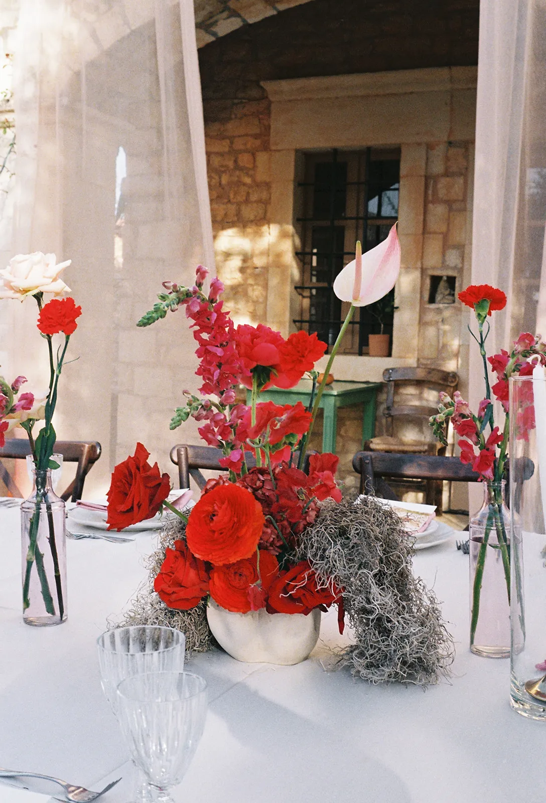 A wedding table centrepiece at Agreco Farm in Greece, with a dense cluster of red garden roses, ranunculus and deep crimson blooms, set off by sprays of pink snapdragons and a single pale pink anthurium rising above. Soft grey-green moss trails down the sides of a cream vase, and slim glass bottles of white and red carnations and pink snapdragons stand on either side. The table is laid with white linen and cross-back wooden chairs under a sunlit stone wall, giving a warm, rustic and romantic feel.