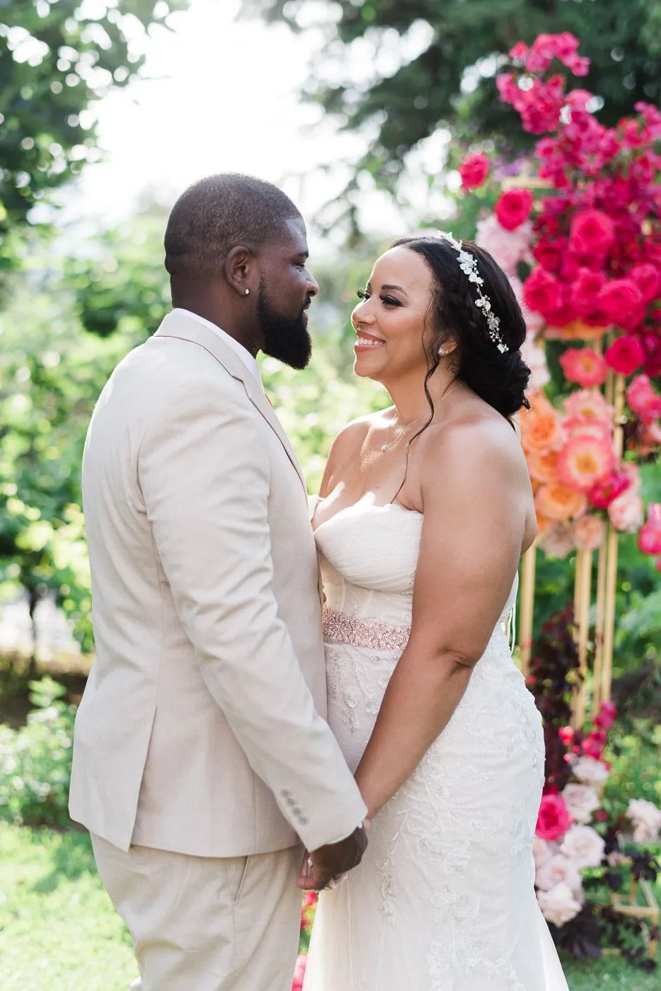 A bride and groom stand face to face holding hands, smiling at each other on a leafy lawn. She wears a strapless lace gown with a jewelled rose-gold sash and a pearl-and-crystal hair vine in her dark updo, while he wears a light beige suit. Behind them, a tall arch of hot pink and coral garden roses and peonies frames the scene in soft, dappled light. These are wedding flowers at Dourakis Winery in Alikampos, Crete, Greece.