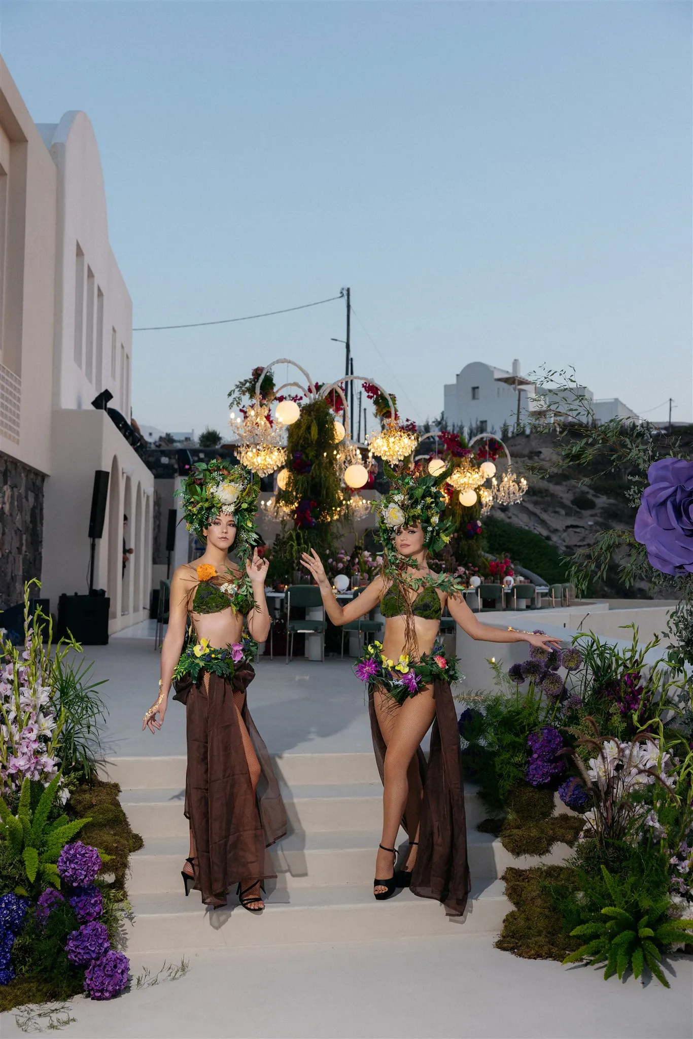 Two models in living floral costumes stand on pale stone steps at Islet Santorini in Thira, Greece, one reaching an arm out toward the other. Their headpieces and bodices are woven from green foliage and white blooms, with flower garlands at the waist over flowing brown skirts. Purple hydrangeas, moss and ferns line the steps, and behind them glowing globe lights hang from arched frames above a table set for a wedding dinner in the early evening.