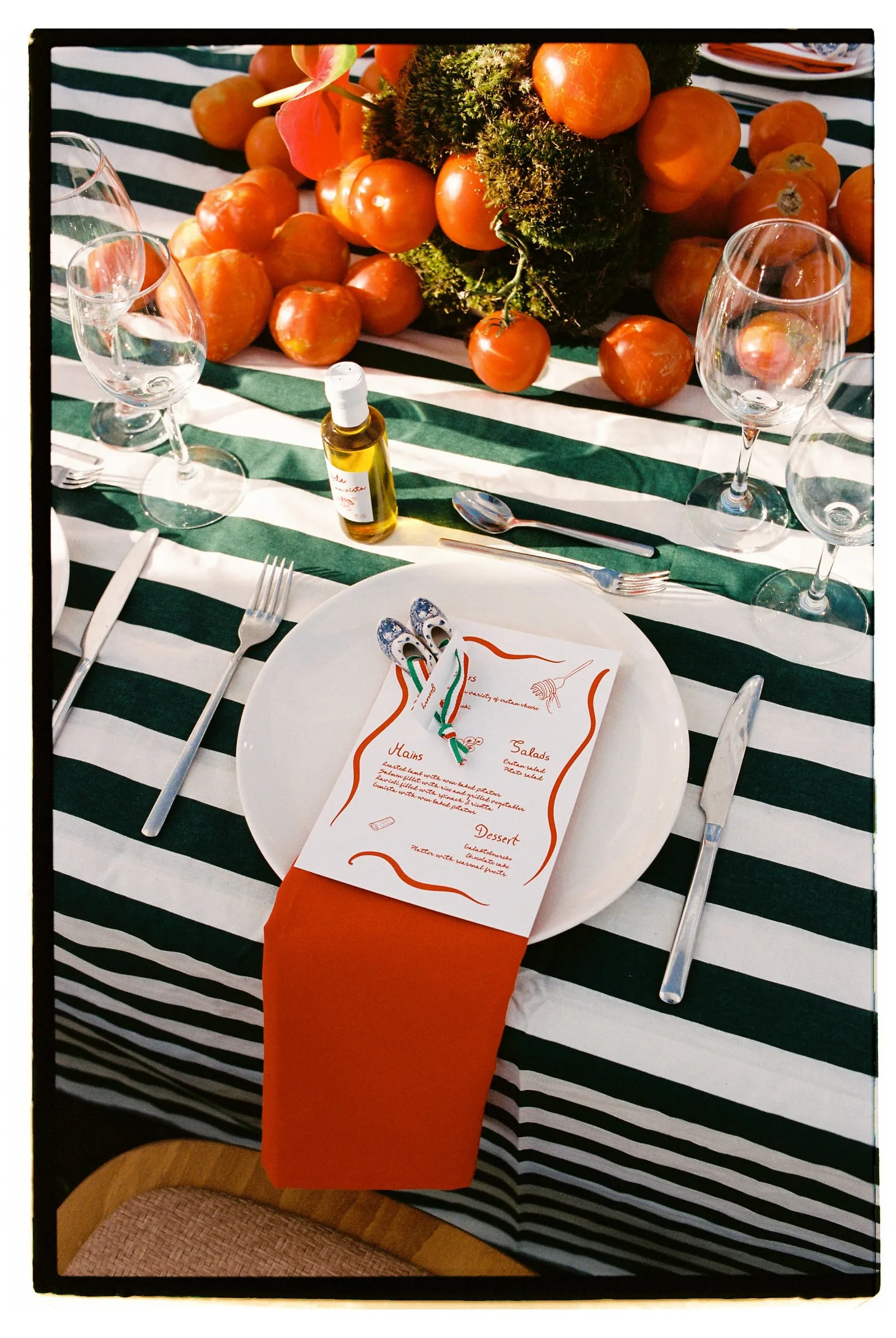 A place setting on a green and white striped tablecloth at Wine Dark Sea Villas in Rodia, Crete, Greece, for a wedding garden ceremony. A white plate holds a menu card with red wavy borders and an orange napkin, topped with two small blue-and-white patterned slippers tied with green and red ribbon. Behind it, a centrepiece of ripe orange tomatoes and green moss sits beside a small bottle of golden olive oil and clear wine glasses.