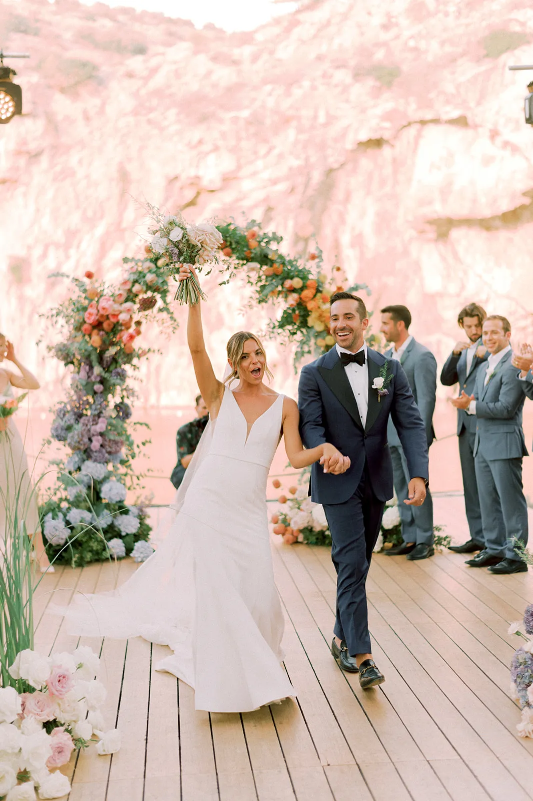 A bride in a white V-neck gown raises a cream and blush bouquet high as she walks along a timber deck beside her groom in a navy suit and black bow tie, both beaming. Behind them stands a curving arch of peach roses, coral blooms and blue hydrangeas, with groomsmen in grey-blue suits clapping on the right. This wedding flower installation is at Lake Vouliagmeni in Greece, with soft pink light glowing across the water behind.