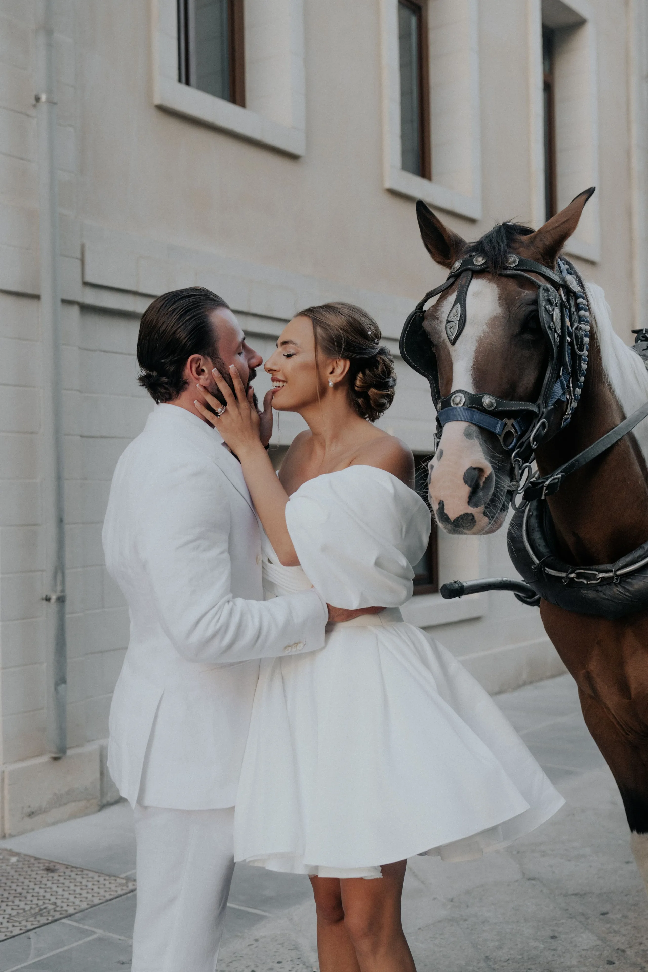 A bride in an off-the-shoulder white gown with a sculpted sleeve and a soft updo holds her groom's face in her hands as they share a tender moment, both smiling, while a brown horse with a white blaze and a dark ornate bridle stands close beside them against a pale stone wall. This is a wedding photograph taken in Greece, with no flower arrangements visible in the frame.
