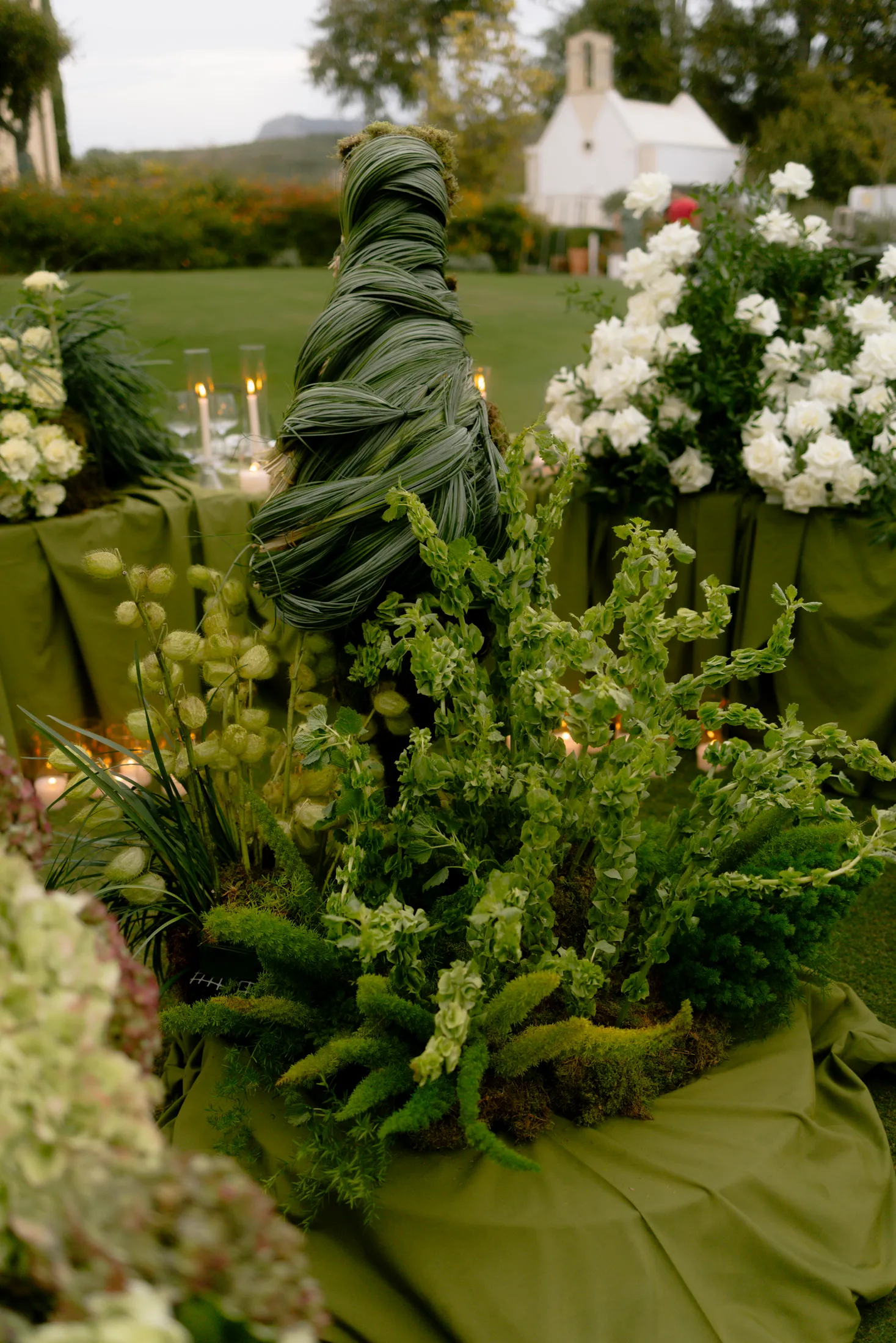 A tall, spiralling column of broad green leaves rises from a low, lush arrangement of trailing green foliage, moss and honesty seed pods on an olive-green draped table, with white roses in a tall vase to the right. These wedding table flowers at Ktima Roxani Estate in Crete, Greece, sit outdoors on a manicured lawn, with a small white chapel and soft evening candlelight in the background.