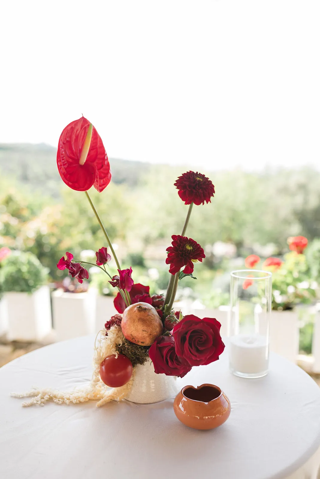 A wedding table centrepiece in deep red at Agreco Farm in Greece, built around tall red anthurium and dark crimson dahlias rising from a small cream vase. Lower down sit red garden roses, a pomegranate, a red tomato, a strawberry and a tuft of pale dried grass, all on a round white tablecloth. A clear glass candle holder and a terracotta dish sit beside it, with soft-focus green foliage and orange blooms behind.