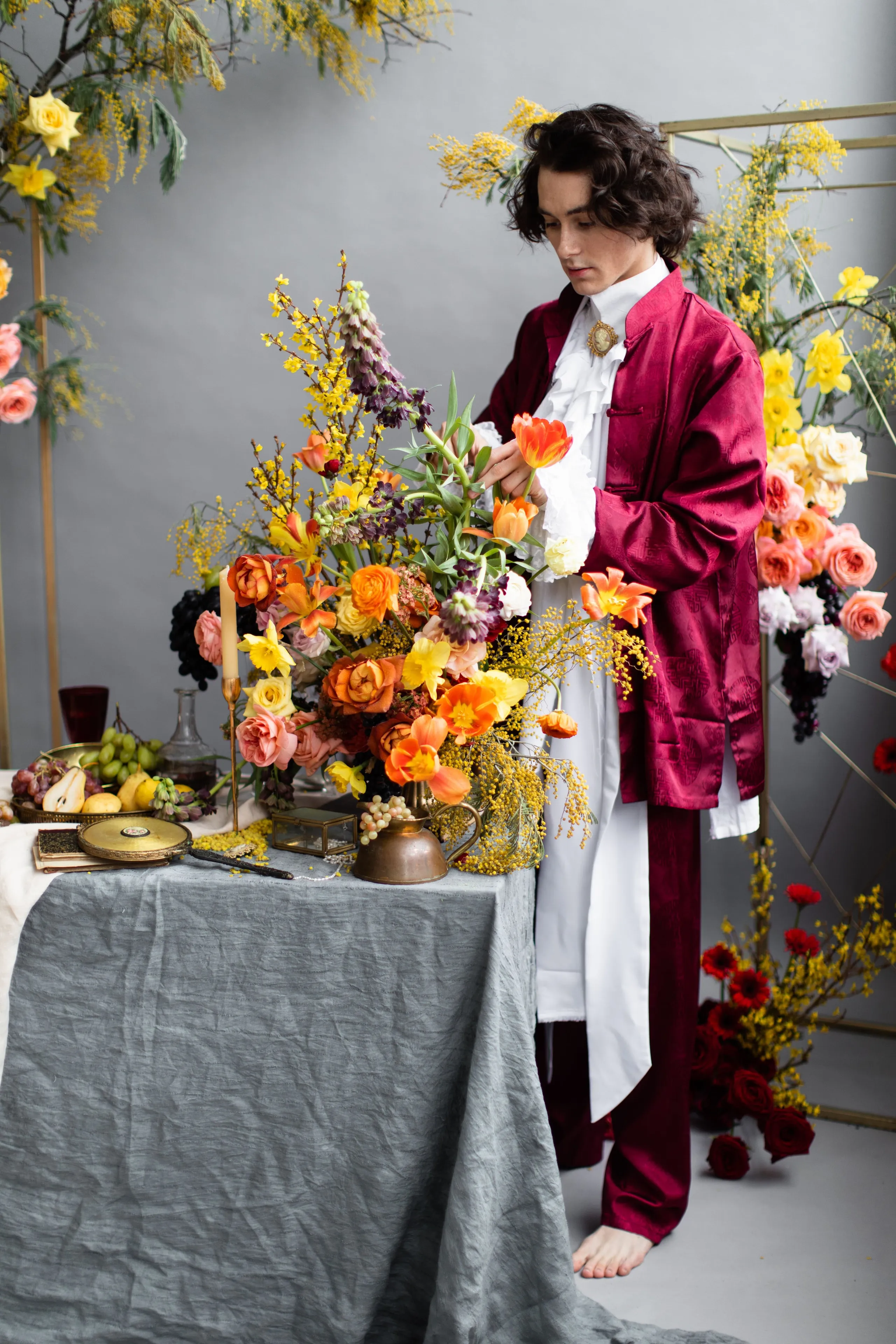 A styled wedding table in Russia, with a tall, lavish centrepiece of orange and yellow tulips, coral and peach garden roses, golden daffodils and feathery mimosa rising from a copper pitcher on a grey linen cloth. Beside it sit a brass candlestick, a bowl of pears and grapes, and a glass decanter, while a person in a deep crimson brocade coat and white ruffled shirt reaches into the arrangement. Behind them, arches of roses and yellow blooms frame the scene in soft grey light, evoking a medieval feast.