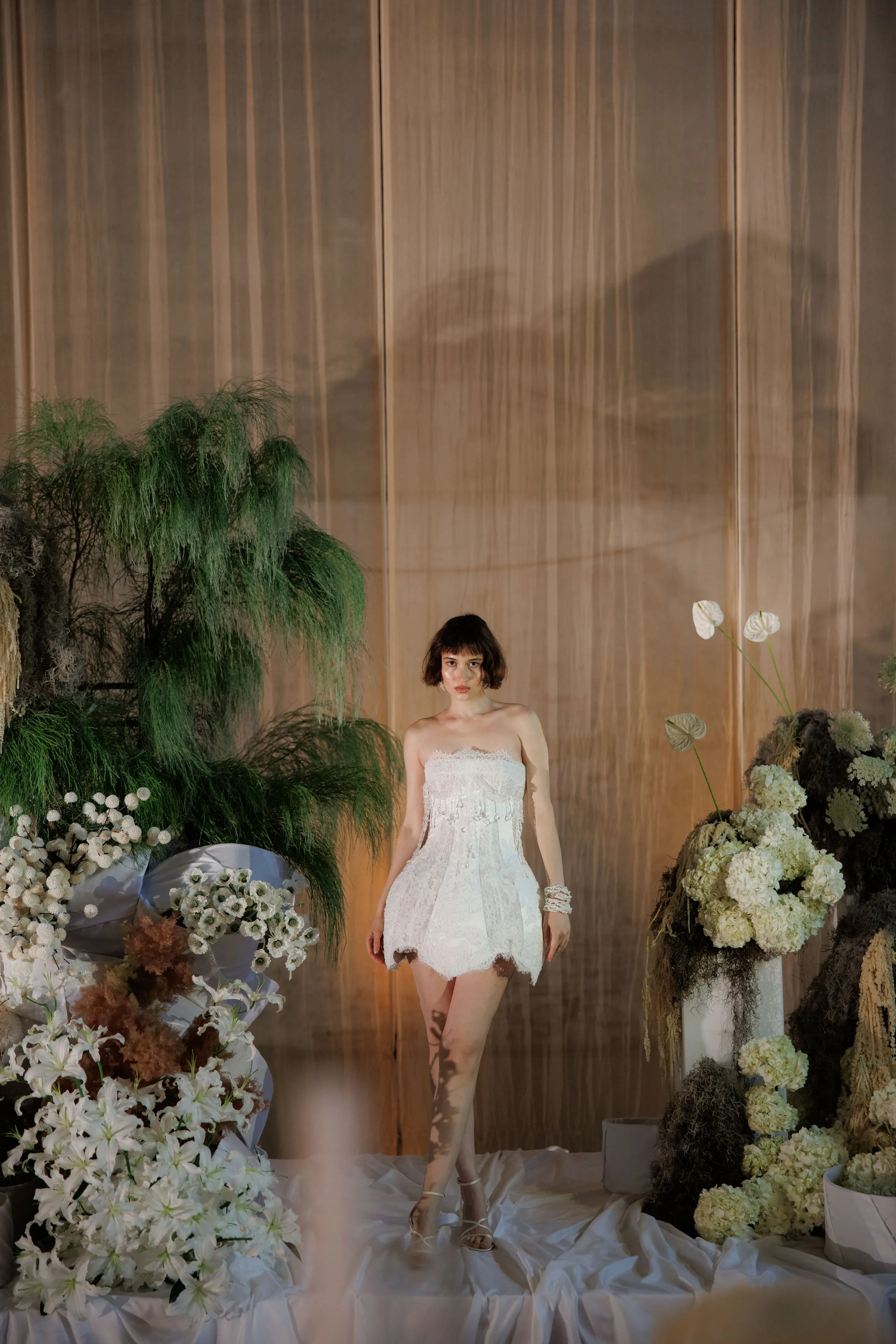 A woman with a short dark bob stands centrally in a strapless white lace mini dress with a beaded bodice, on a white silk-draped stage in Rethimno, Crete, Greece. Around her, wedding flowers in creamy whites and greens fill the scene: white lilies in a low arrangement at left, tall feathery asparagus fern behind her, and white hydrangeas and anthurium blooms rising from the right. Soft, warm light falls on sheer gauzy drapes behind her, giving the scene a quiet, elegant mood.