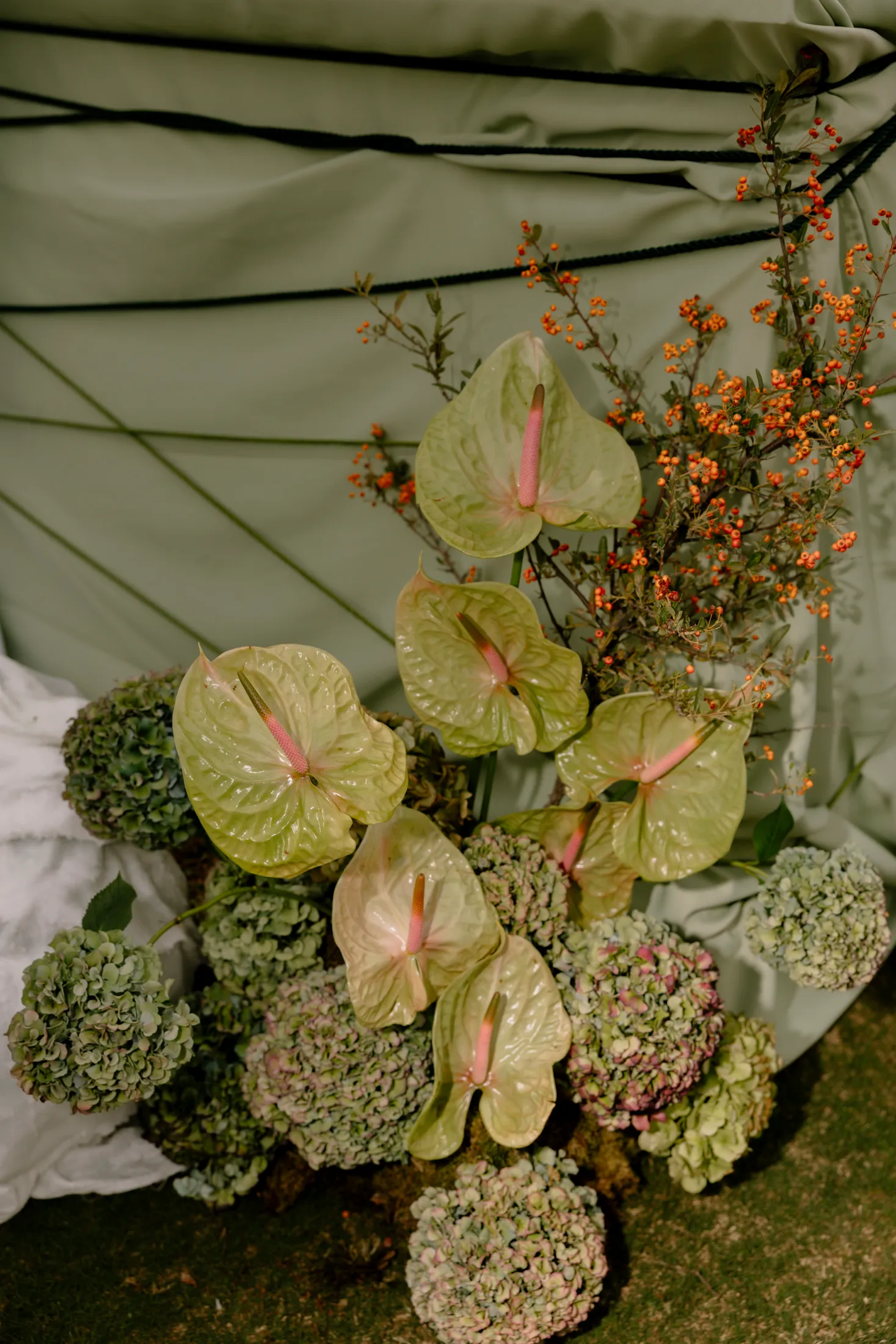 Pale green anthurium blooms with pink spadices rise from a low cluster of green and blush hydrangea heads, with sprigs of orange berries tucked behind them. The arrangement sits on a moss-covered floor against a sage-green drape with dark cords, forming a wedding floral installation at Ktima Roxani Estate in Armenoi, Crete, Greece.