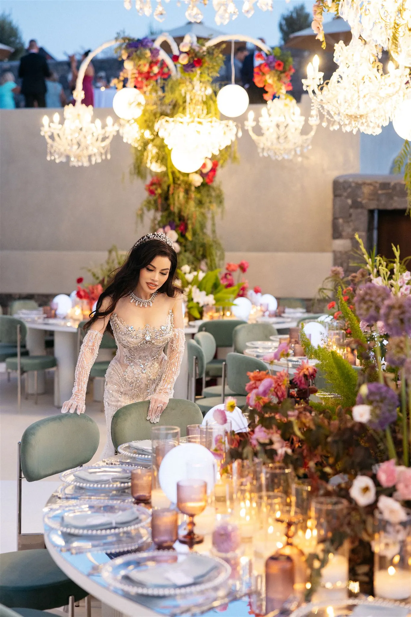 A bride in a sparkling, beaded gown with long sheer gloves and a jewelled tiara leans over a green velvet chair at a candlelit wedding table on a terrace at Islet in Santorini, Greece. Below her, a long table is laid with pearl-rimmed glass plates and amber glasses, and tall arrangements of purple alliums, pink and white roses and warm orange blooms spill along its length. Crystal chandeliers and glowing white orbs hang above a floral arch, and the soft dusk light gives the scene a glamorous, quietly theatrical mood.