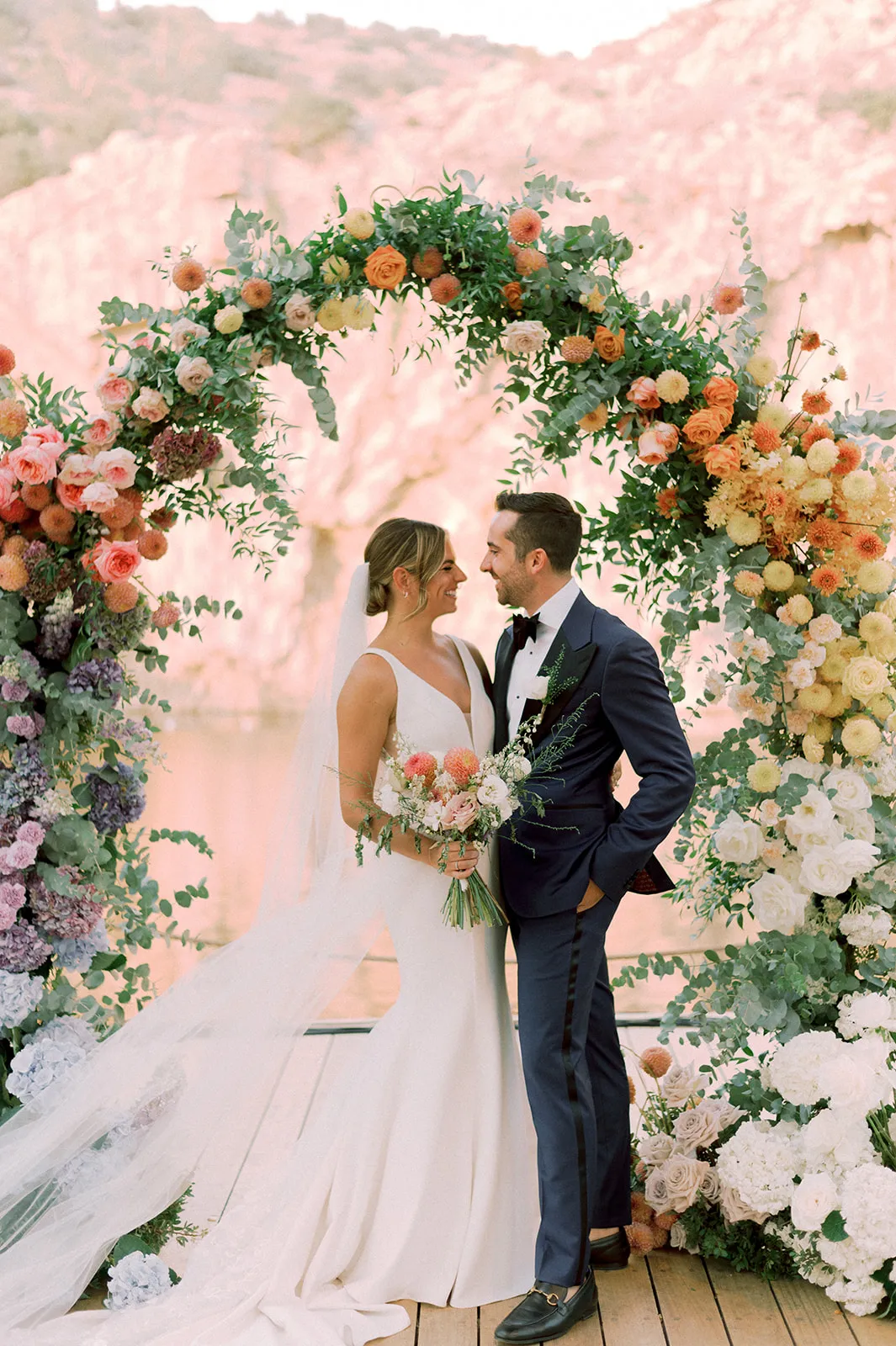 Bride and groom face each other, smiling, beneath a tall floral arch at a wedding venue at Lake Vouliagmeni in Greece. The arch is dense with garden roses, orange and cream dahlias, white roses, lilac hydrangeas and trailing eucalyptus, shading from blush pink through peach to deep violet. The bride, in a white satin gown with a long veil, holds a bouquet of pink and peach blooms, and the groom wears a navy suit with a black bow tie. Soft, warm light and a blurred pink backdrop give the scene a dreamy, romantic feel.