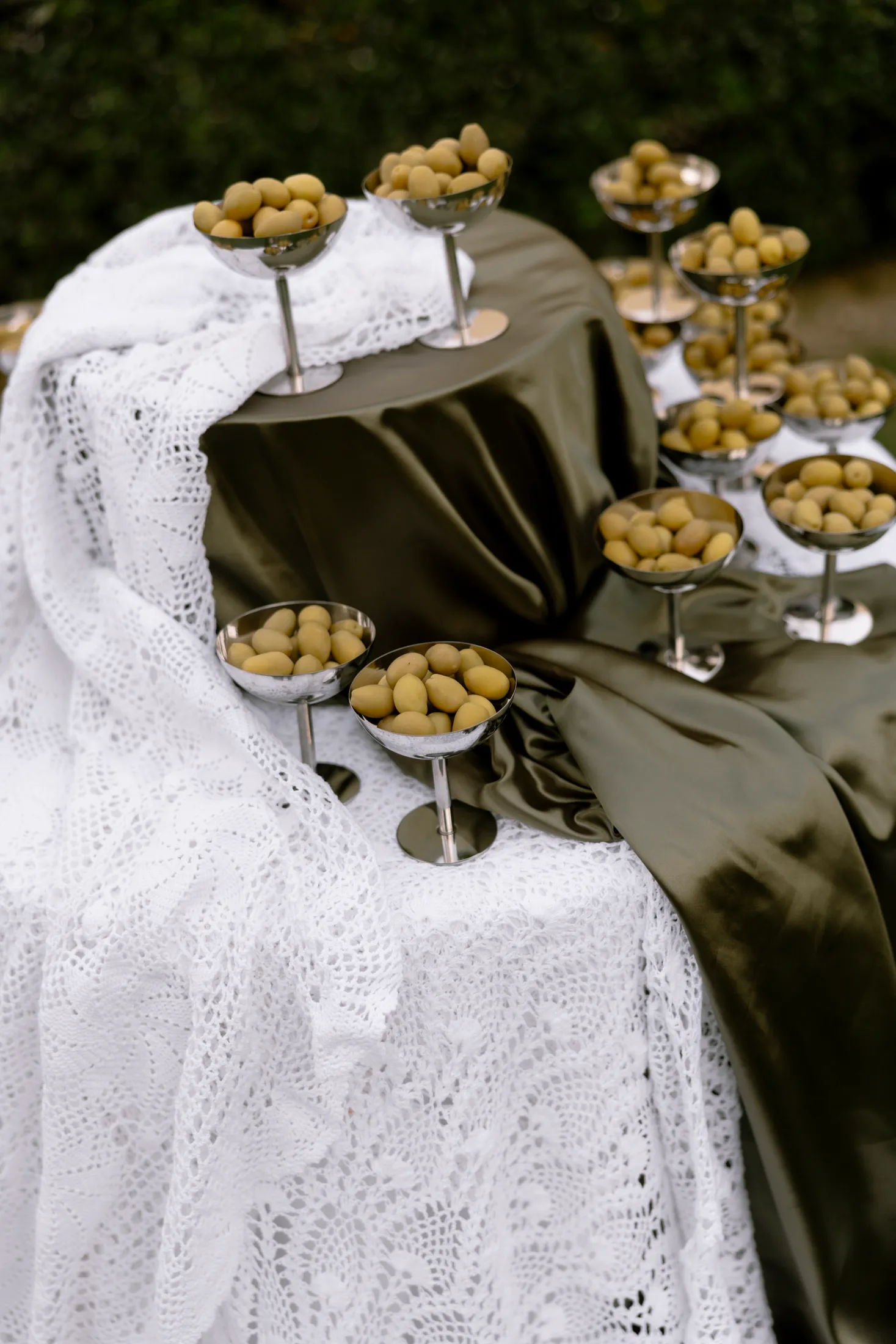 Silver footed coupe glasses, each filled with small green olives, are arranged along a table draped in dark olive-green satin and a white crocheted lace cloth that spills over the edge. Dense green hedging forms the blurred backdrop, and soft daylight falls across the scene. This is a wedding table styling at Ktima Roxani Estate in Armenoi, Crete, Greece, with the olive theme echoing the island's landscape.