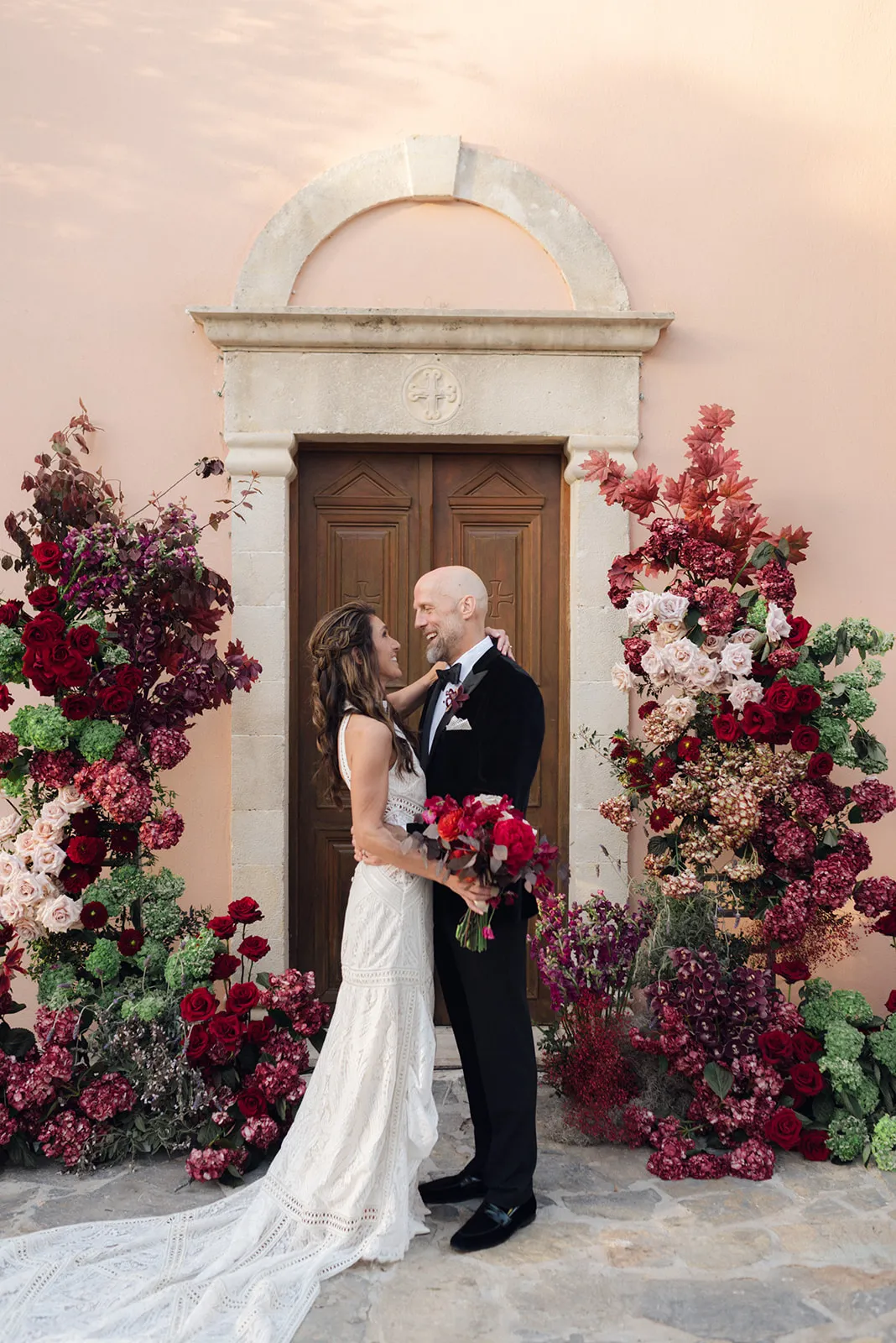 A bride in a lace-paneled ivory gown and a groom in a black velvet tuxedo embrace and smile at each other in front of a wooden double door, framed by towering floral arches of deep red roses, burgundy hydrangeas, cream roses and green hydrangeas. The bride holds a bouquet of red peonies and dark foliage. This is a wedding flower display at Agreco Farm in Greece, with a warm, romantic mood.