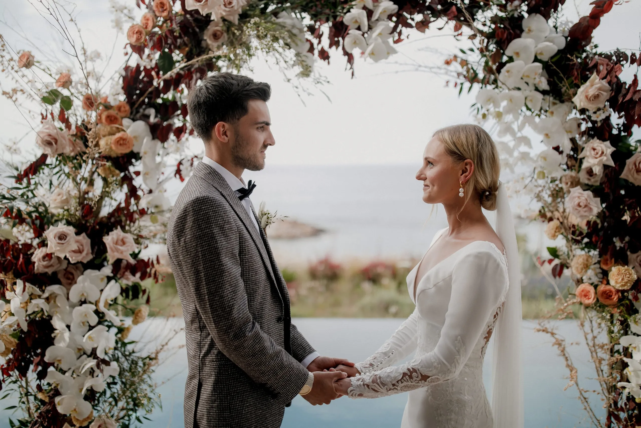 A bride and groom stand facing each other, holding hands beneath a floral arch of peach and blush roses, white orchids and deep burgundy foliage, with the sea glimpsed behind them. He wears a grey check suit and black bow tie, and she has a fitted white gown with lace sleeves, a sleek low bun and a long veil. This is a wedding ceremony in Crete, Greece, at Nuez Villas.