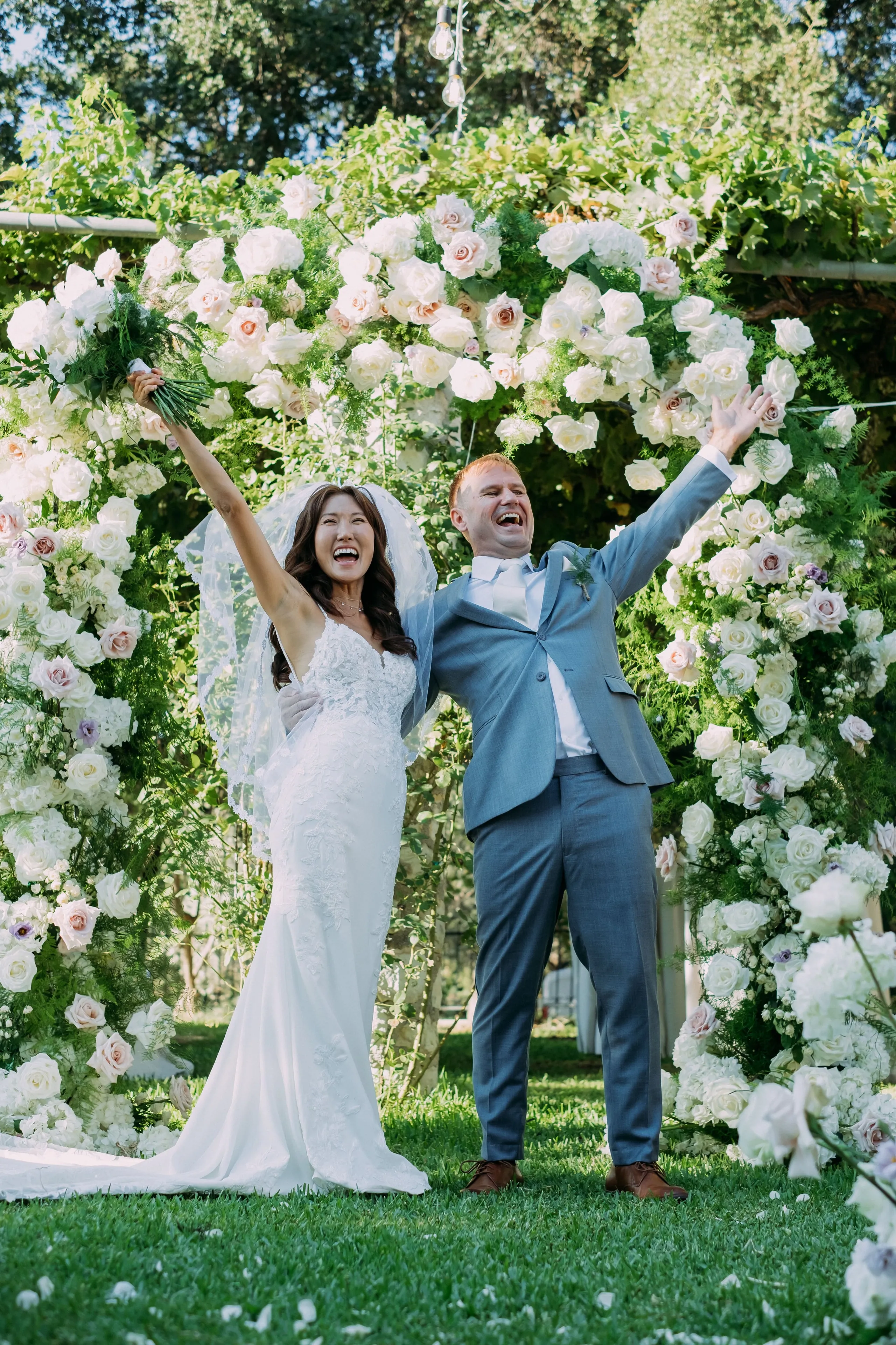 A bride and groom stand laughing with their arms raised in triumph on a lawn, in front of a full circular arch of white and blush garden roses, with trailing greenery and sprigs of fern. The bride, in a fitted lace-detailed gown and veil, holds a green-wrapped bouquet aloft, while the groom wears a dusty-blue suit. This wedding arch and flowers were created by manfler for a wedding at Dourakis Winery in Alikampos, Crete, Greece.