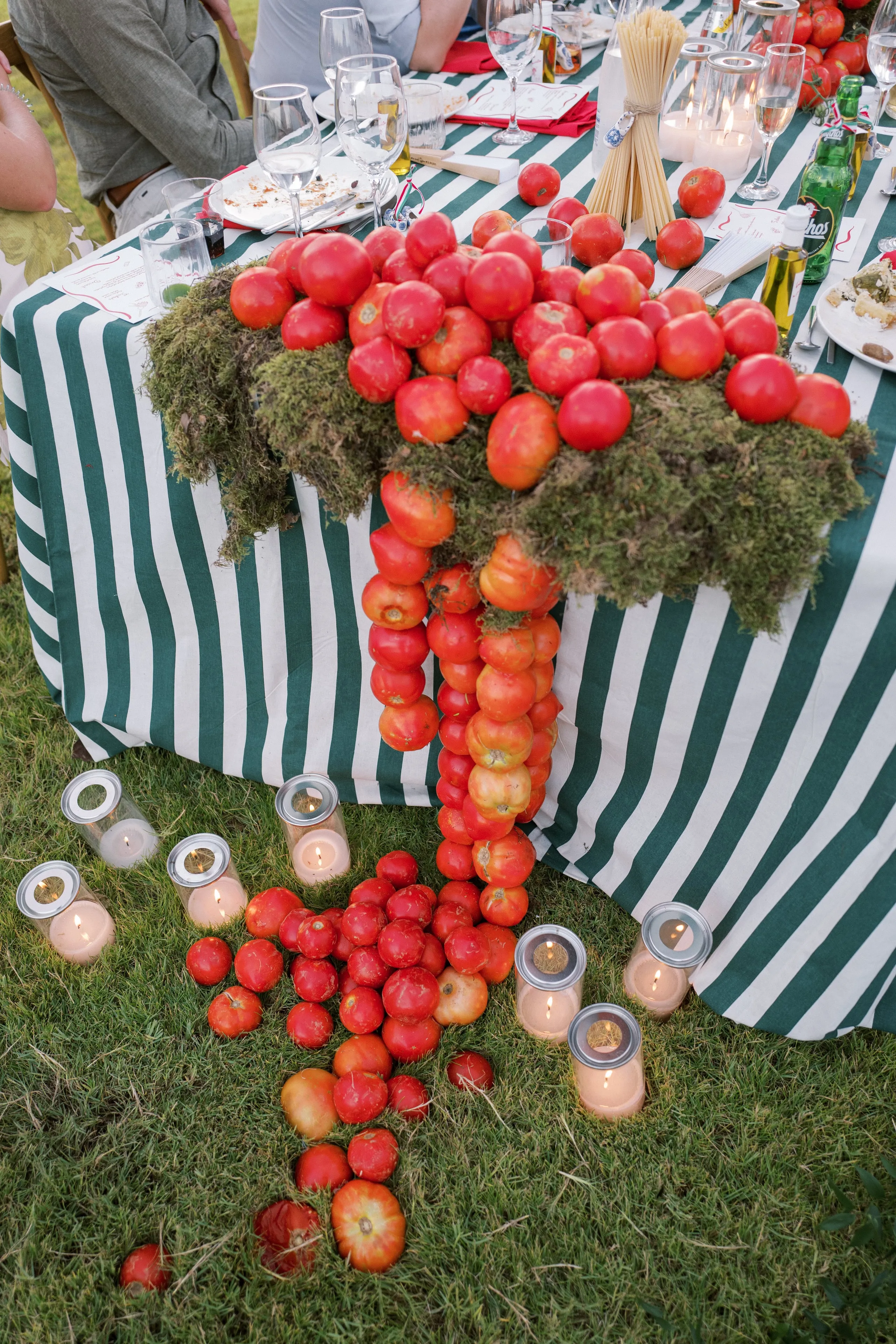 A long garden table dressed in green-and-white striped linen is decorated with cascading red tomatoes, many on the vine-like ripeness of heirloom varieties, spilling over beds of green moss and tumbling down onto the lawn. Glass hurricane candles glow amid the scattered tomatoes at the table's edge, and bundles of dried spaghetti, wine glasses and place settings complete the scene. This event styling is for a garden wedding dinner at Wine Dark Sea Villas in Santorini, Crete, Greece.