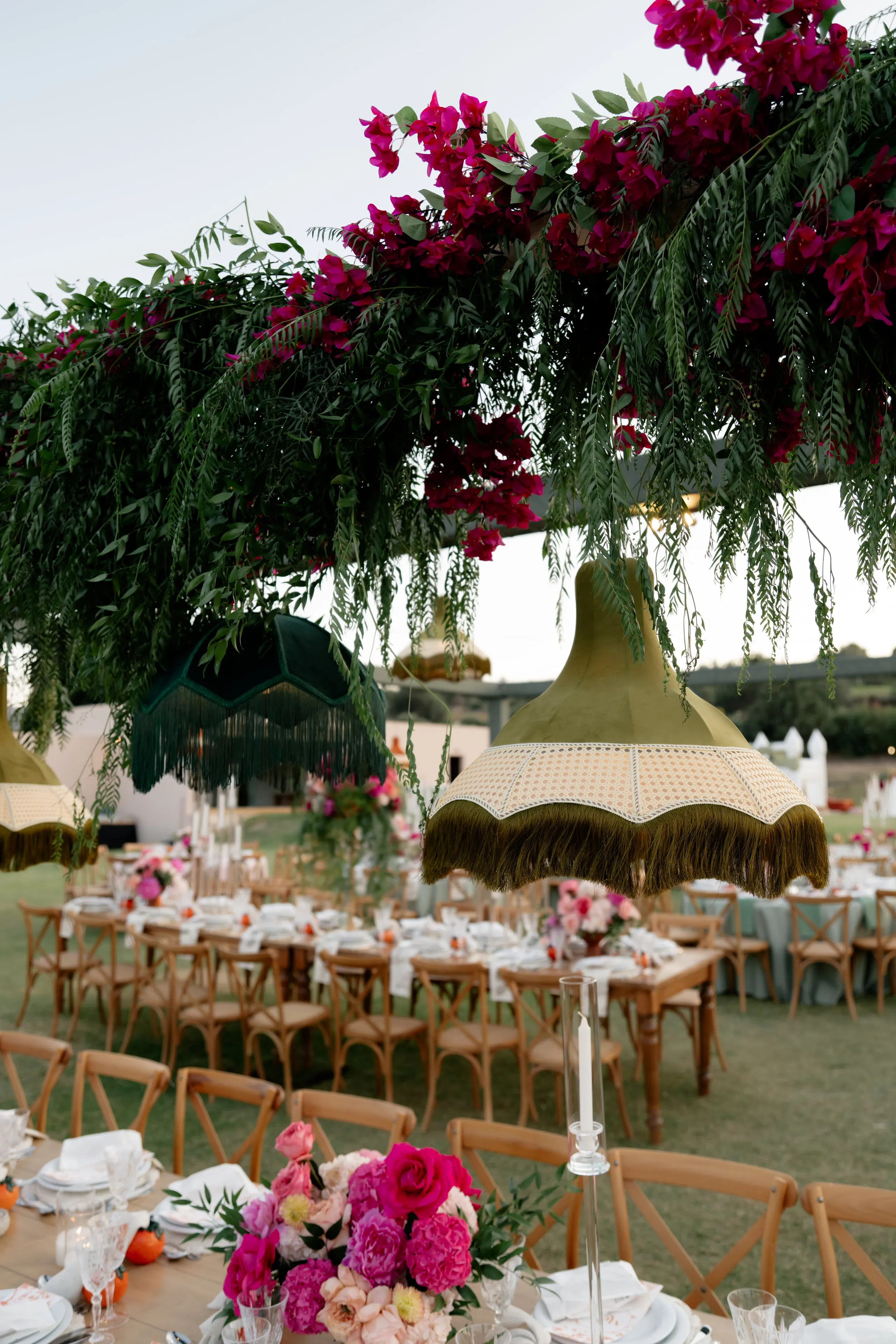 Hanging over a lawn dinner at Crete Golf Club in Chersonisos, Greece, a canopy of trailing green fern-like foliage is studded with magenta bougainvillea, while olive-green fringed lampshades sway beneath it. In the foreground, a low centrepiece of hot pink and blush peonies, roses and ranunculus sits on a long wooden table set with white linens and clear glasses. Rows of cross-back wooden chairs stretch across the grass toward more pink floral arrangements in the soft evening light, all part of a wedding flowers display in Greece.