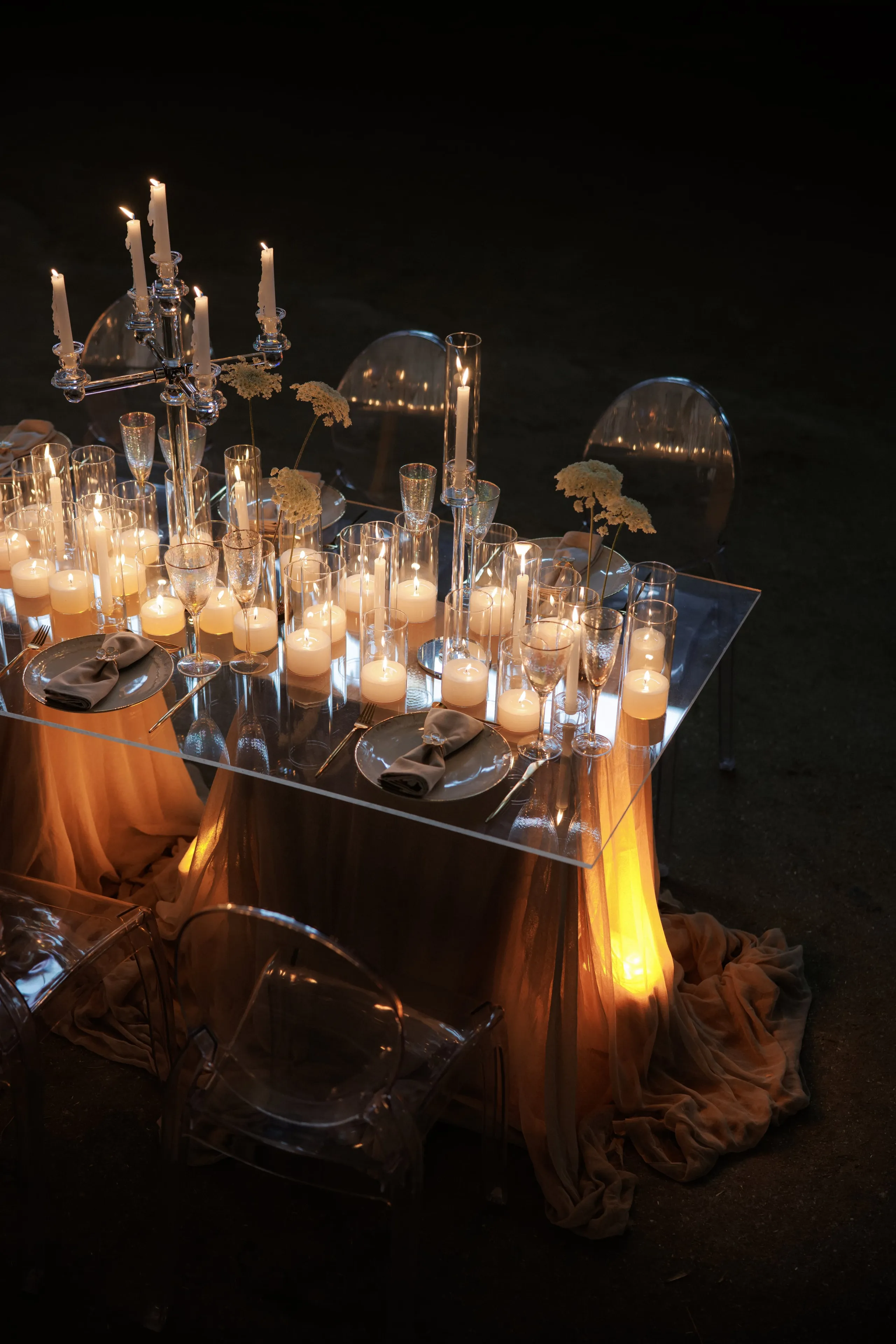A wedding table set at night in Rethimno, Crete, Greece, lit almost entirely by candles. A clear acrylic table sits on the dark ground, draped below with soft peach chiffon that glows amber in the light. Tall glass hurricane votives and taper candles line the table alongside stemless glassware, grey-rimmed plates with folded grey napkins, and sprigs of small cream wildflowers, while a crystal candelabra rises at the back. Clear ghost chairs complete the intimate, romantic scene.
