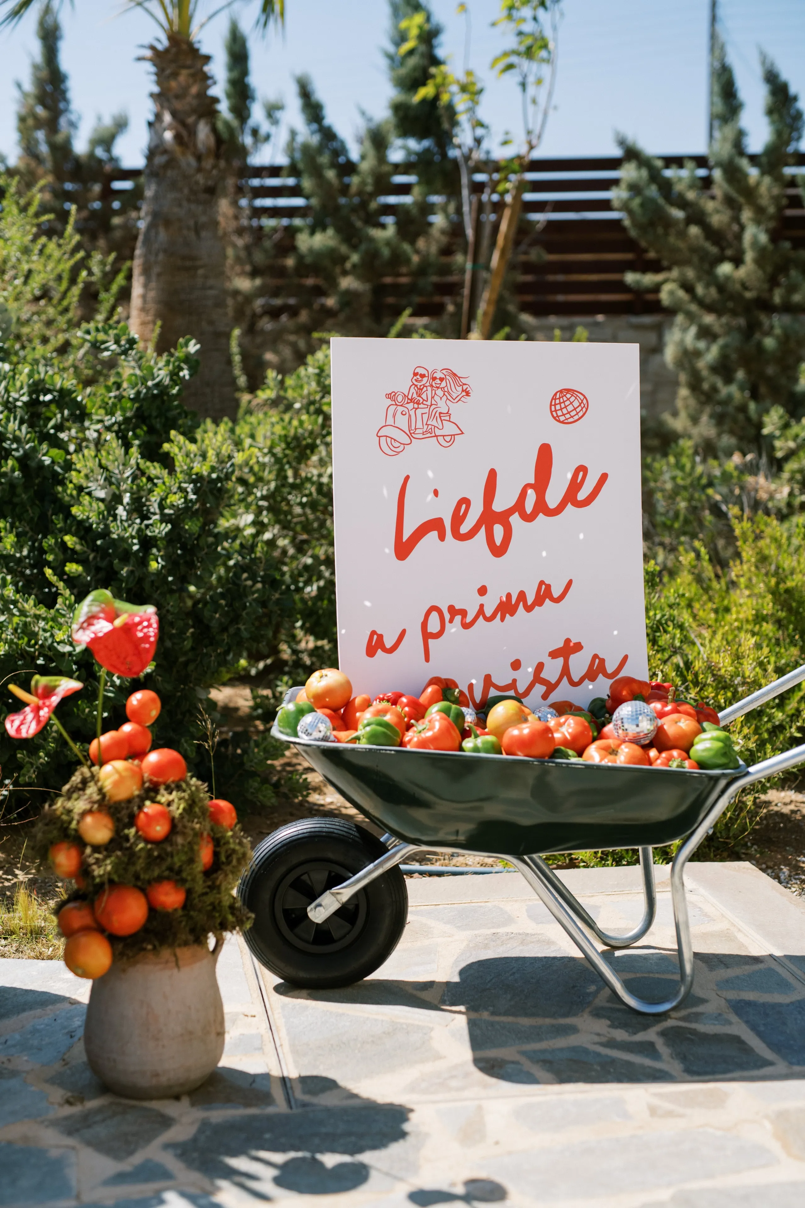 A metal wheelbarrow brimming with ripe red and green peppers, tomatoes and small disco balls sits on a stone-paved courtyard in front of a white sign with red hand-lettered text reading "Liefde a prima vista" beside a small red scooter illustration. To its left stands a rustic clay pot of red anthuriums and tomatoes on moss, in a garden of green shrubs and palms. This wedding decor is at Wine Dark Sea Villas in Rodia, Crete, Greece, bright and playful in the midday sun.
