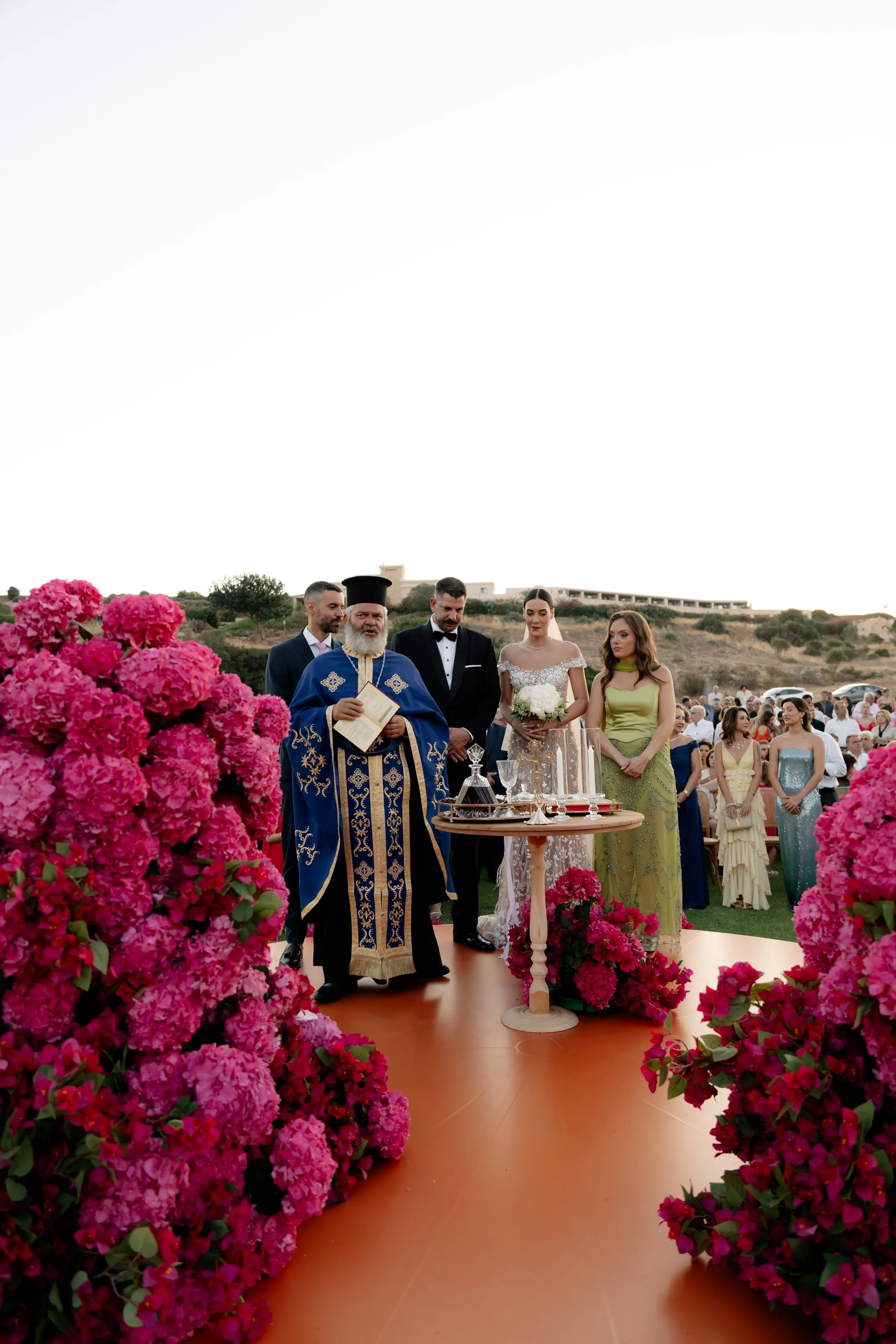 Pink hydrangeas and magenta bougainvillea spill in tall, sculptural masses along both sides of a terracotta-coloured aisle, leading toward a wedding ceremony at Crete Golf Club in Chersonisos, Greece. A bearded Orthodox priest in a royal blue vestment edged with gold stands at a small wooden table set with a wine carafe and candles, while a bride in an off-the-shoulder lace gown holds a white rose bouquet beside a groom in a dark tuxedo. Bridesmaids in green and pale blue gowns stand behind them under a wide, pale sky, with guests and distant hillside buildings beyond.