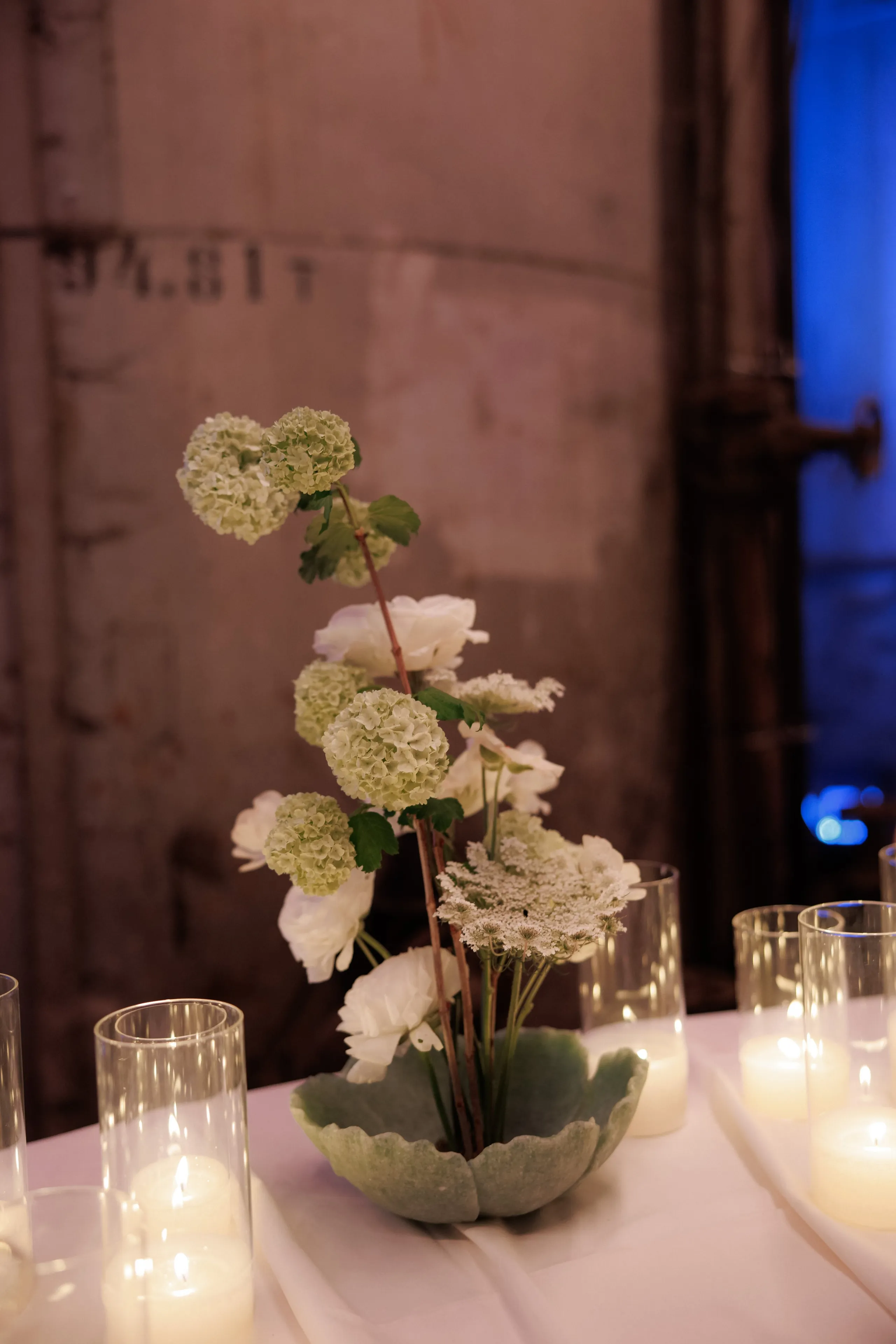 A tall, airy table centrepiece of white wedding flowers rises from a sage-green scalloped ceramic bowl: green-tinged snowball blooms, white ranunculus and lacy white Queen Anne's lace on slender branching stems with small green leaves. It sits on a white-draped table in Rethimno, Crete, Greece, flanked by clear glass candle holders with soft flames, against a weathered grey concrete wall with a blue light glowing at the edge.
