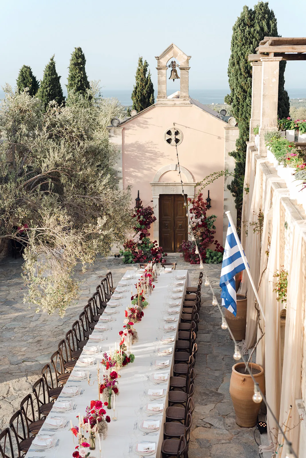 A long white-clothed wedding table set beneath an old olive tree in a stone courtyard at Agreco Farm in Greece, with a pale pink chapel behind it. Deep burgundy and red blooms, likely garden roses and foliage, are clustered in tall arrangements at the chapel doors and run down the centre of the table among tapered candles. String lights hang above, and a Greek flag flies from a balcony at right beneath soft, late-afternoon light.
