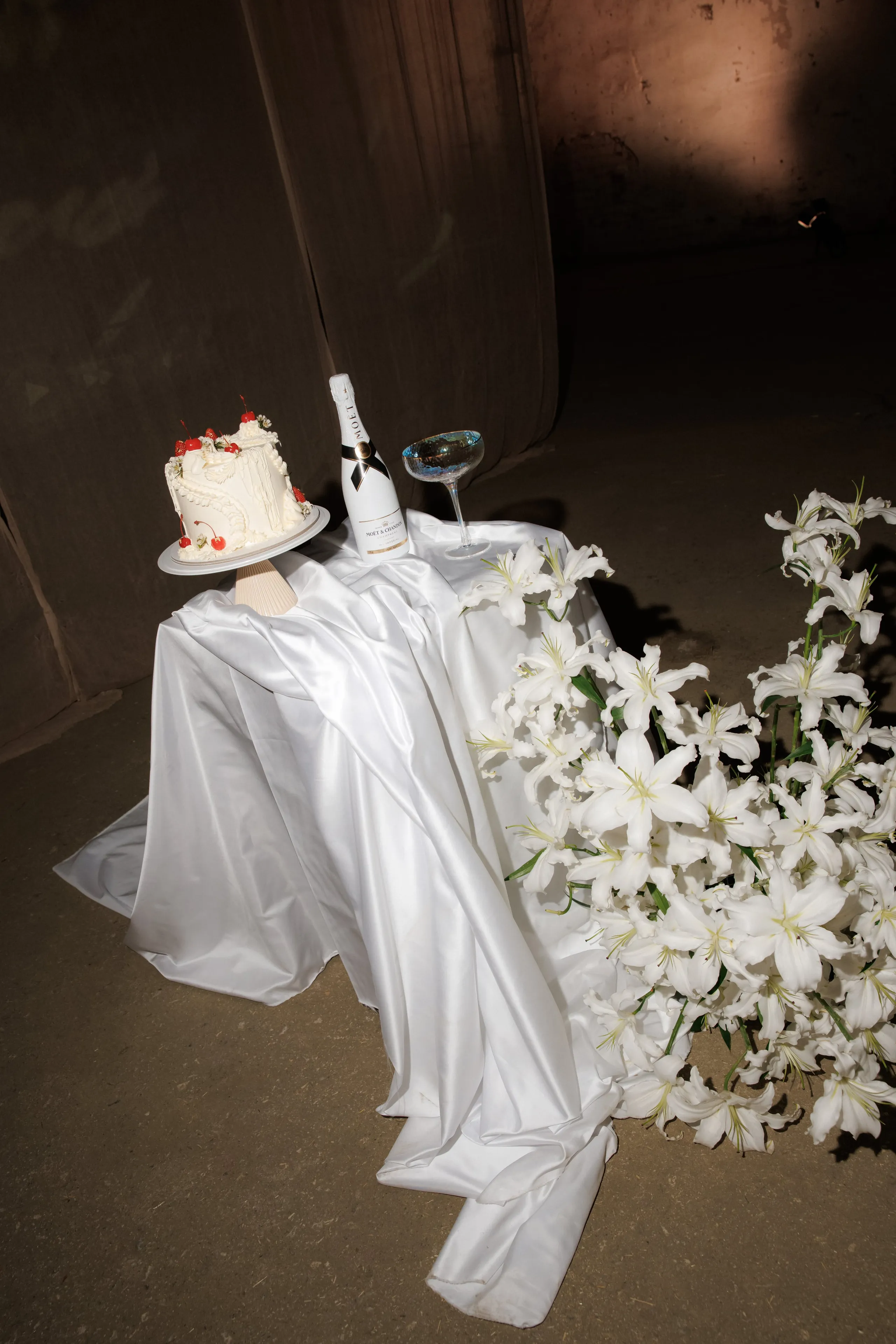 A tumble of white lilies spills across the stone floor at the right, their trumpet-shaped blooms and green stems draped over a crumpled white satin cloth. On the cloth sit a small white-iced cake topped with red cherries, a white bottle of Moët champagne, and a shallow glass with a blue-tinted rim, all in soft, moody light. This wedding flower display in Rethimno, Crete, Greece, has a dramatic, minimalist feel against the dim walls.