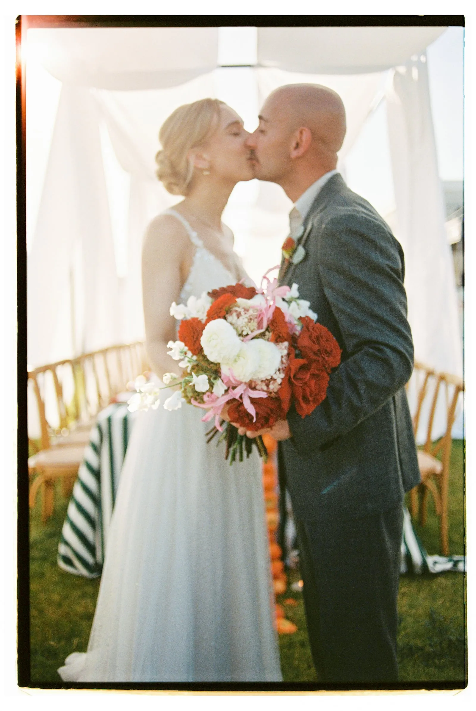 A bride and groom share a kiss beneath a white draped canopy on a lawn at Wine Dark Sea Villas in Rodia, Crete, Greece. She holds a bridal bouquet of red garden roses, white ranunculus, pink lily blooms and sprigs of white sweet peas, while he wears a grey suit and a small boutonniere. Behind them stand wooden chairs and a striped green-and-white table, all bathed in soft, warm sunlight.