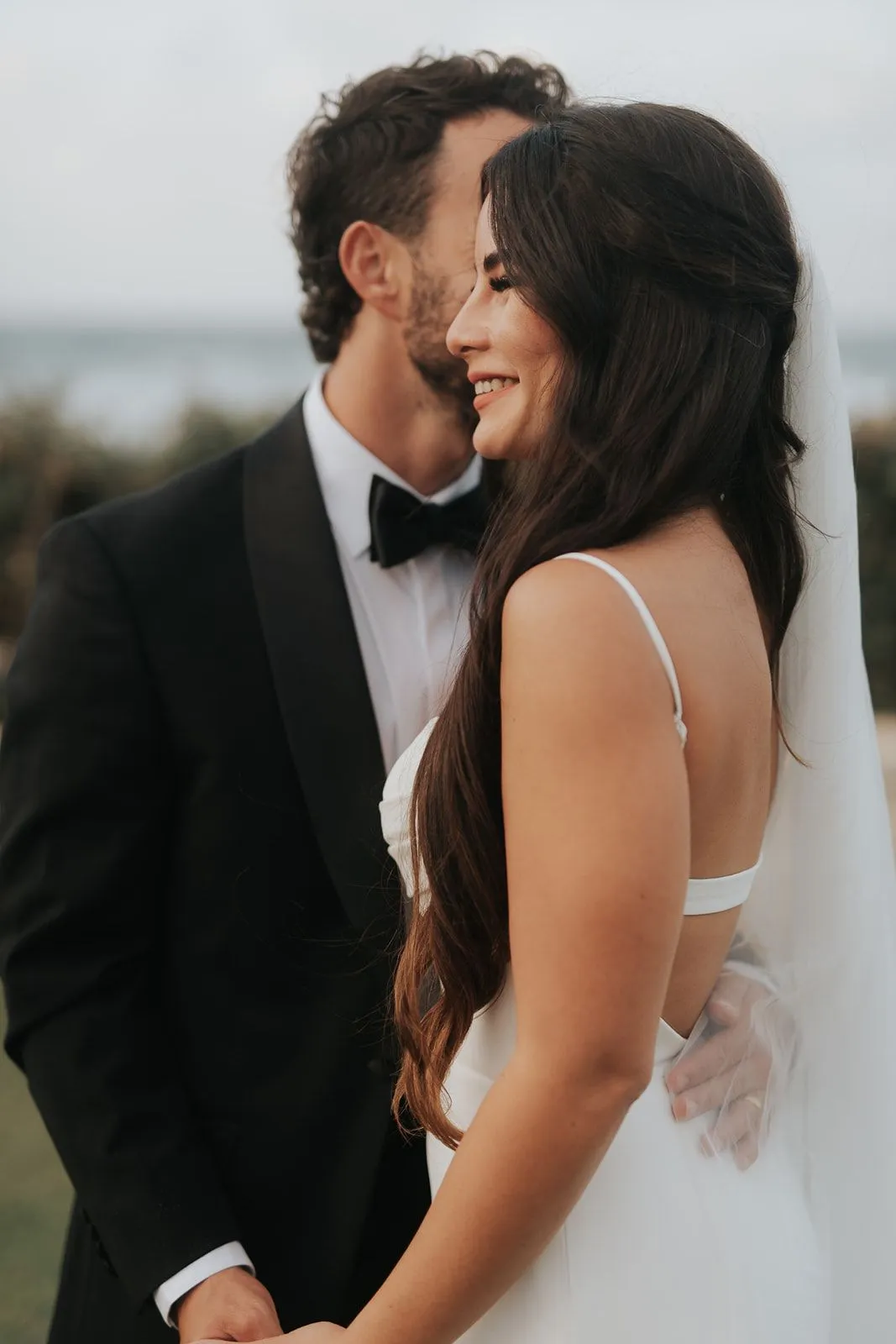 A close, soft-focus portrait of a bride and groom standing side by side, the groom in a black tuxedo with a black bow tie and white shirt, his dark curly hair and stubble close to the bride's head as he leans in. The bride, with long dark wavy hair and a sheer veil falling behind her, smiles warmly with her face turned away from the camera toward the sea. Behind them the sky is pale and hazy with blurred greenery at the edges, a wedding portrait taken in Greece.