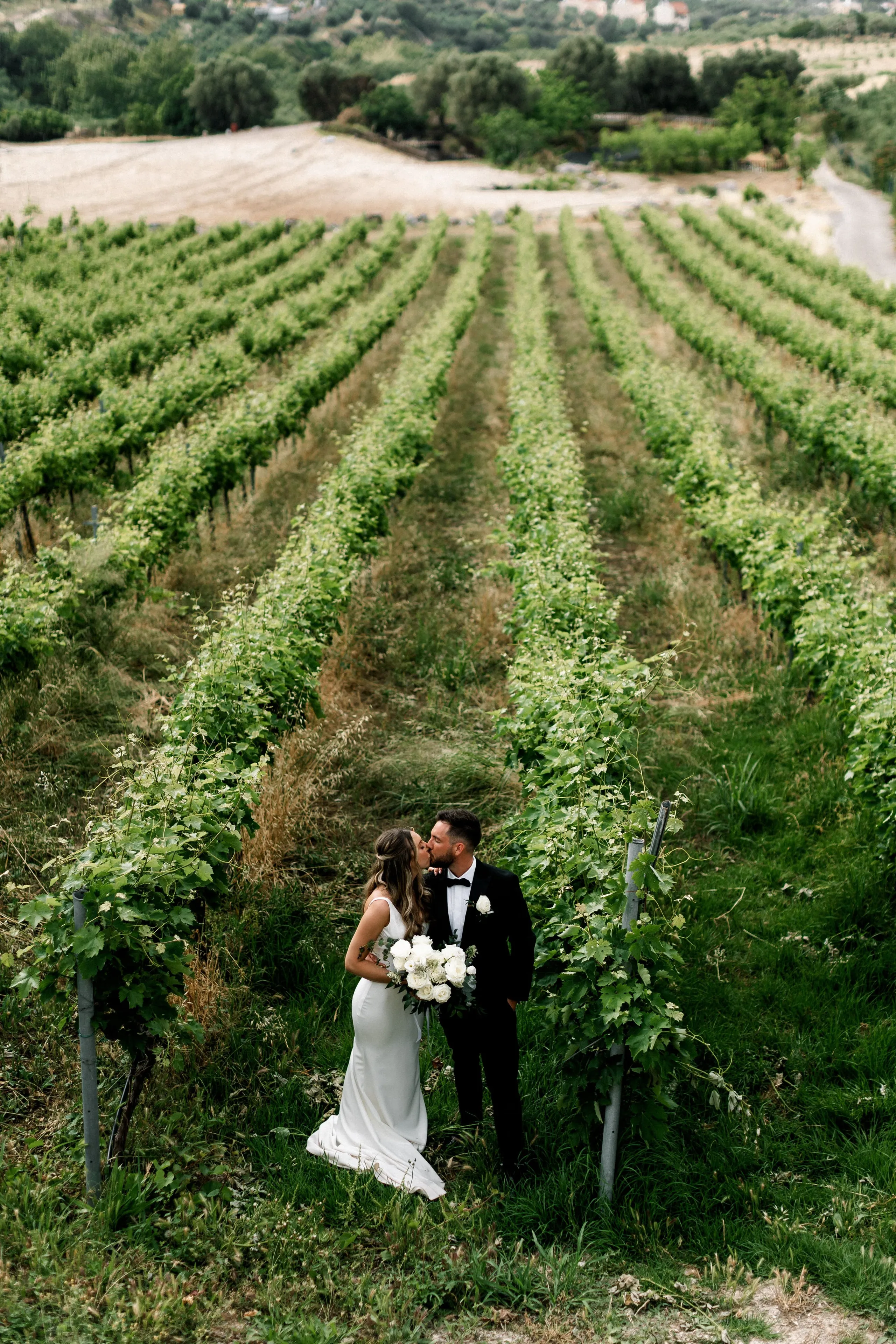 A bride in a fitted white satin gown and a groom in a black suit with bow tie share a kiss between rows of leafy green vines at AgrecoFarms in Adele, Crete, Greece. She holds a full bridal bouquet of white garden roses and hydrangea-like blooms, with a single white flower pinned to his lapel. Behind them the vineyard rows sweep toward a hillside of olive trees and village houses under soft, overcast light.