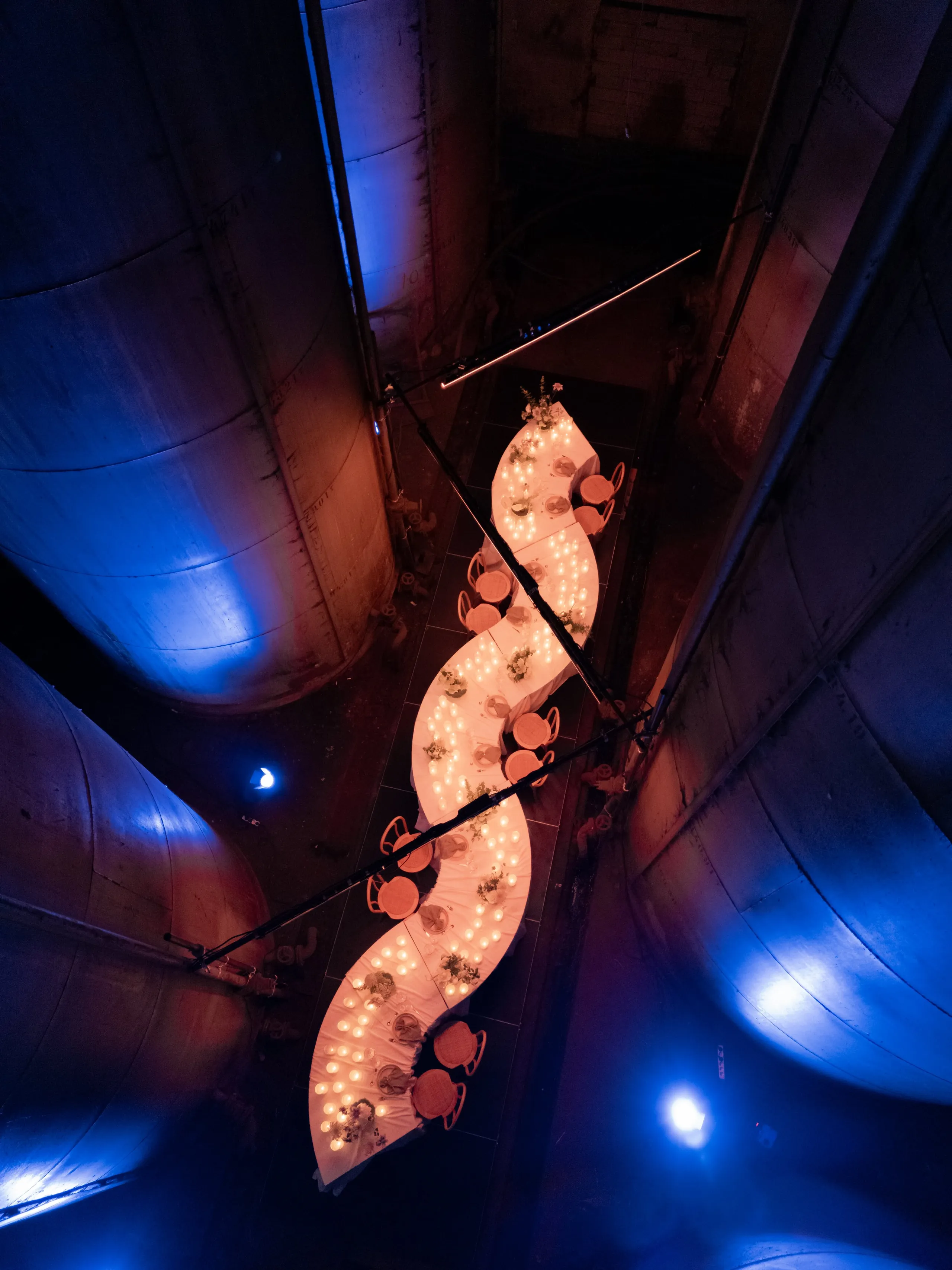 A long, winding banquet table draped in white cloth runs down the centre of a vast industrial space, its S-curve lined with rows of glowing candles and small sprigs of white-green foliage at intervals. Orange chairs line both sides, and the whole scene is bathed in deep blue light from above that contrasts with the warm amber of the candlelight. This wedding table setting in Rethimno, Crete, Greece, is photographed from high above, with the curved concrete walls of a former industrial tank rising on either side.