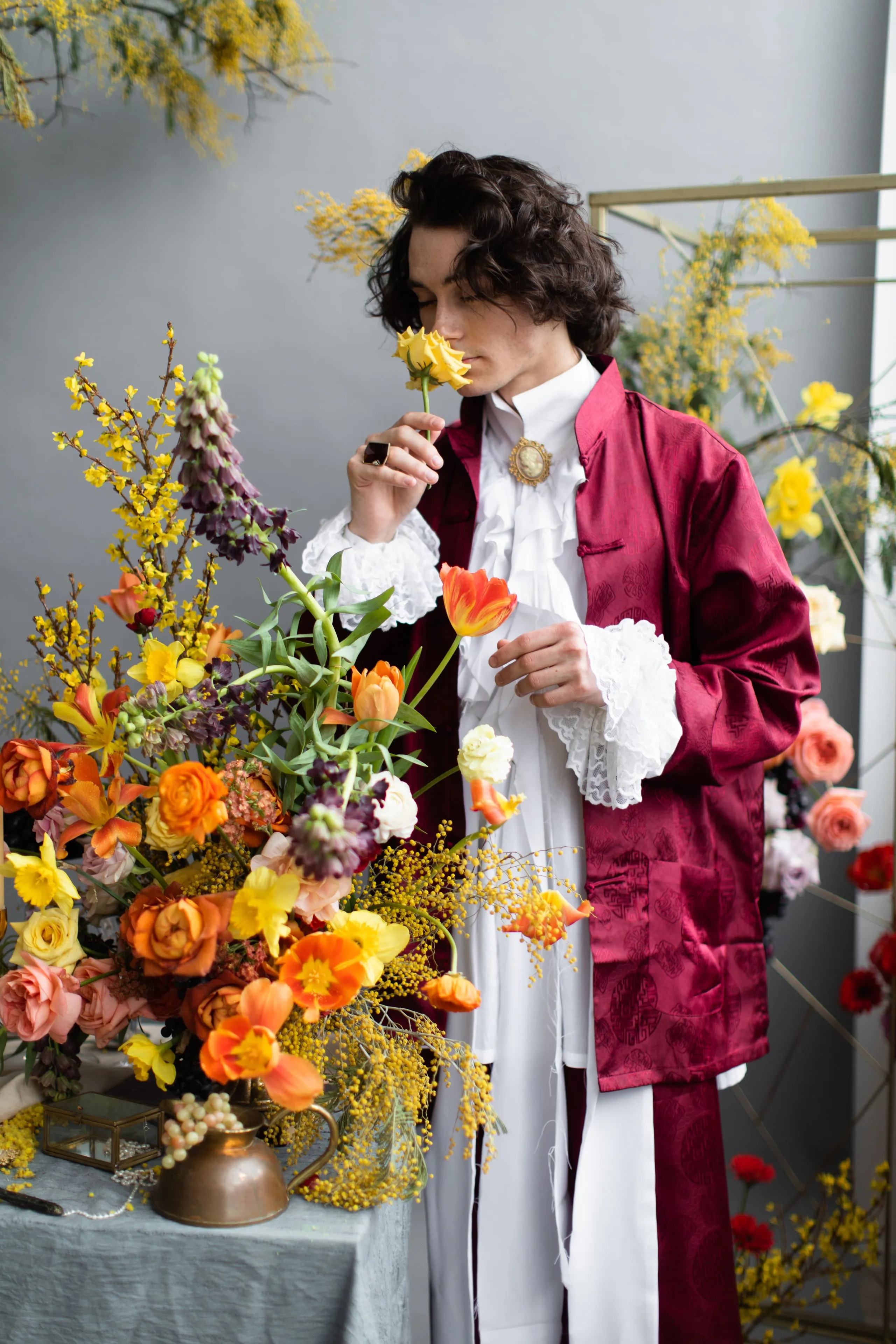 A man in a deep burgundy silk jacket and a white ruffled shirt with lace cuffs stands beside a tall, tumbling arrangement of orange tulips, yellow daffodils, peach garden roses, forsythia and mimosa, with purple foxglove spikes rising above. He holds a single tulip near his face, and a cameo brooch sits at his throat. This event flower styling for a medieval-inspired dinner was created by a florist in Russia.