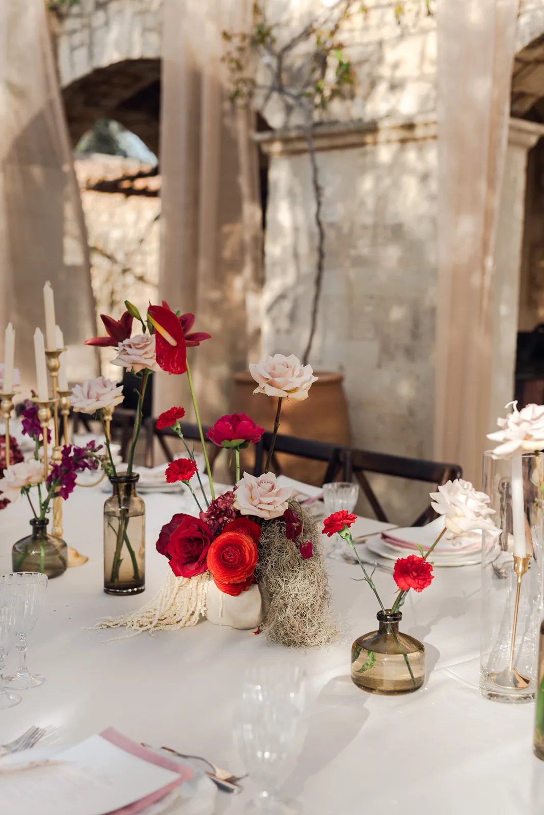 A wedding table set with a low, lush centrepiece of red ranunculus, deep red roses, blush garden roses, red anthurium and carnations, softened with trailing Spanish moss, while slender bud vases of single stems and purple stock stand along the white tablecloth. Gold candlesticks with white tapers and clear glassware catch the warm, dappled light of the stone walls at Agreco Farm in Greece, with a terracotta urn in the background. This is a wedding table flower design for the Where Nature Meets Forever story.