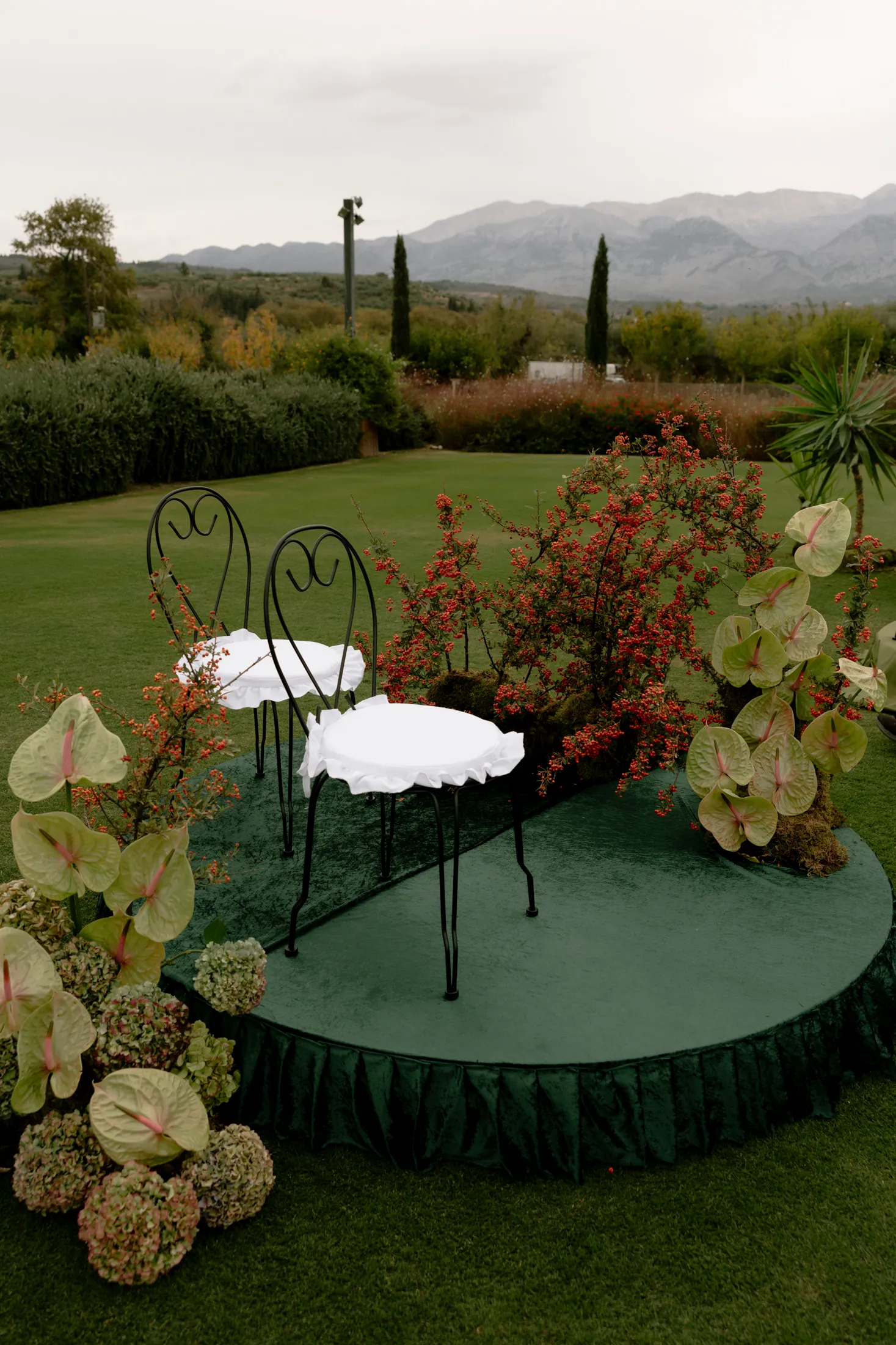A curved green velvet platform on a lawn holds two black wrought-iron chairs and a small round table with white ruffled cloths, set among trailing red-berried shrub, pale green anthurium and blush hydrangeas. Behind is a clipped hedge, cypress trees and a grey mountain ridge under an overcast sky. This wedding flower installation at Ktima Roxani Estate in Crete, Greece is calm, green and understated.