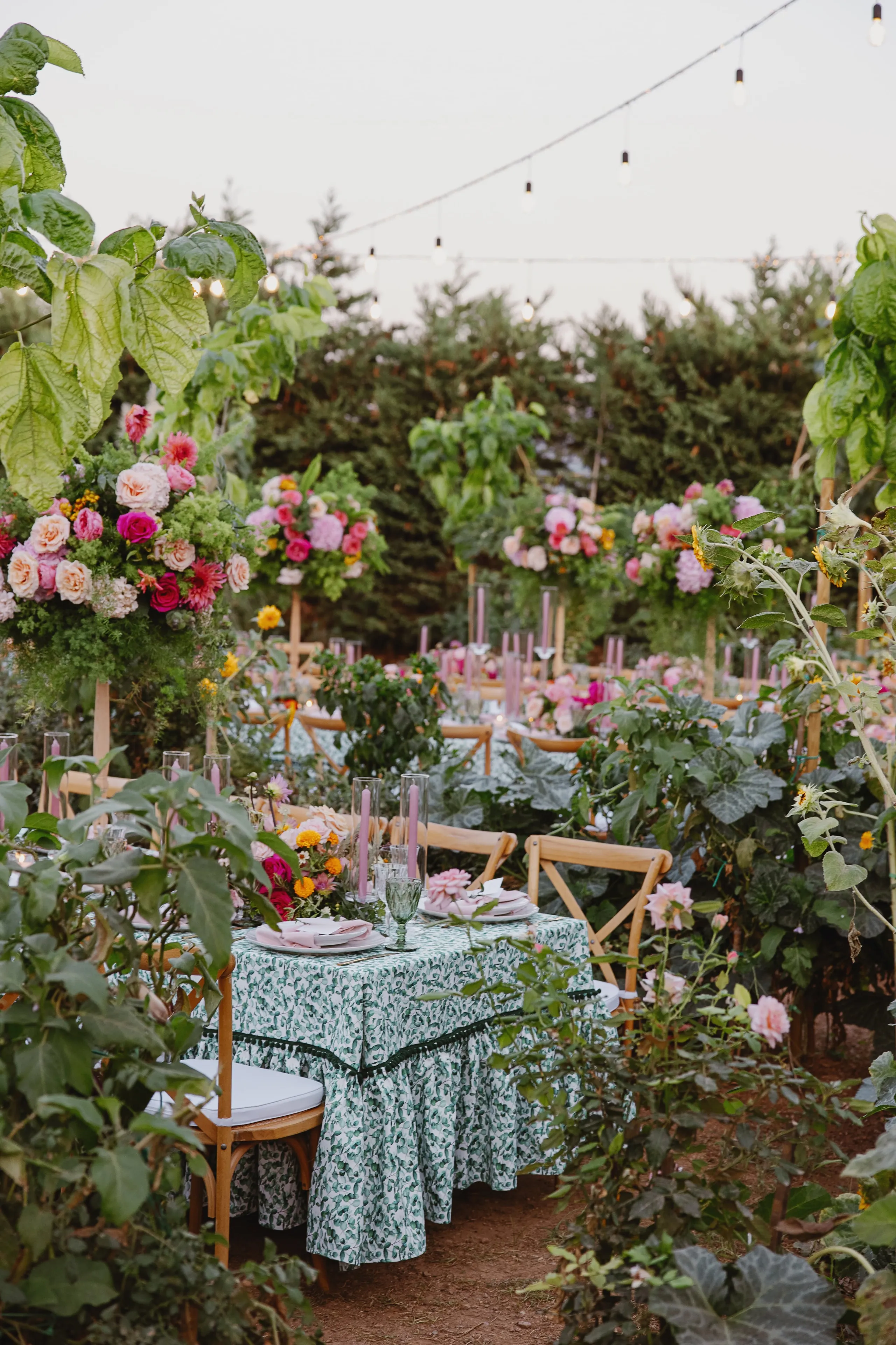 A wedding table setting in a lush garden in Greece, with a green and white patterned cloth, pink plates and lilac candles in glass hurricanes. Behind it, tall floral arrangements of pink roses, peonies and dahlias on wooden stands rise among leafy green plants and sunflowers, with rows of cross-back chairs stretching into the greenery.
