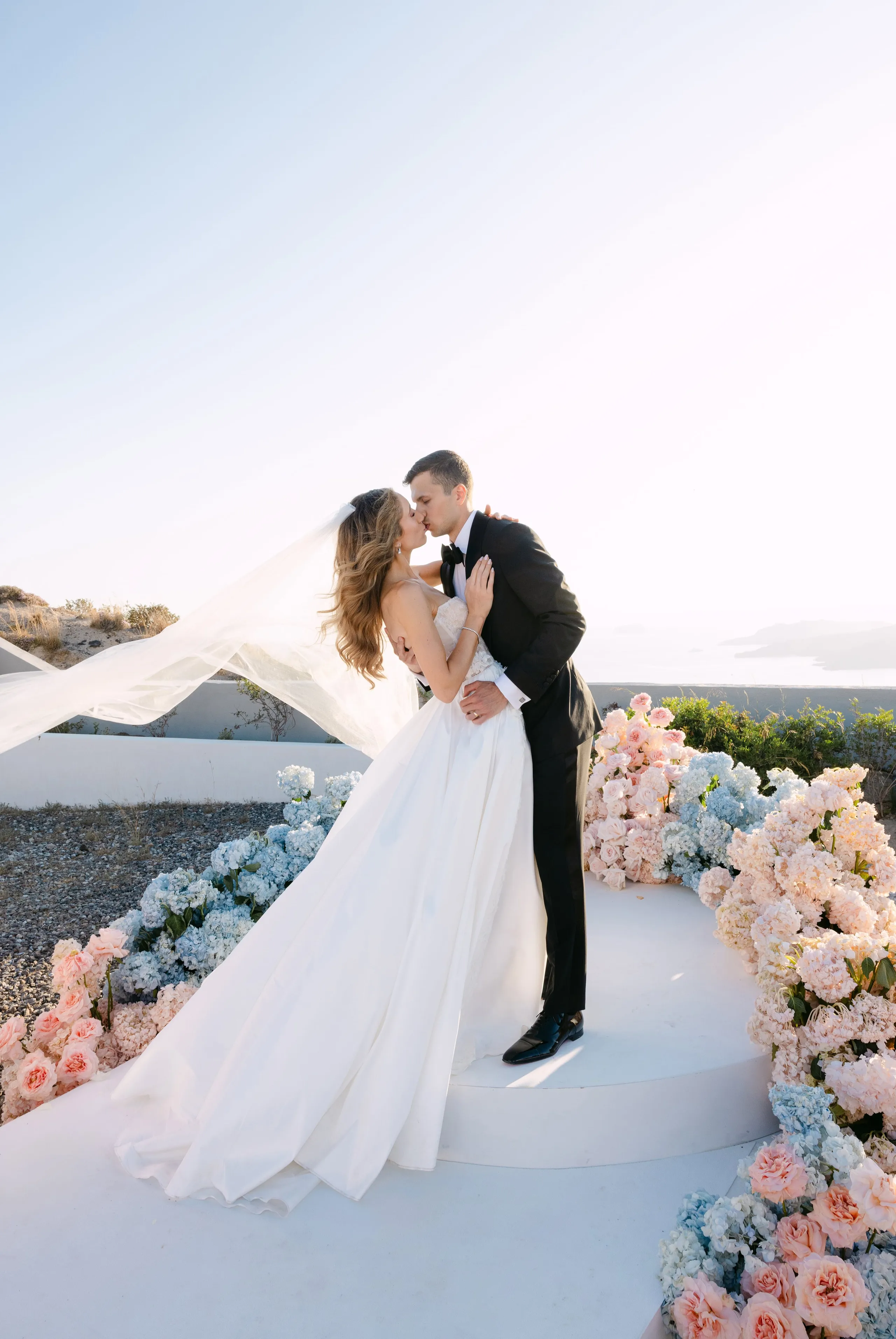 A bride in a strapless white gown and a groom in a black tuxedo with bow tie share a kiss on a white circular platform, her long veil streaming behind her in the breeze. Along the edges of the platform and the path beside it, pale blue hydrangeas and soft pink garden roses spill in lush arrangements, part of a wedding flower installation at Eros Santorini Boutique in Thira, Santorini, Greece. Bright, hazy sunlight and an open sky frame the romantic scene.