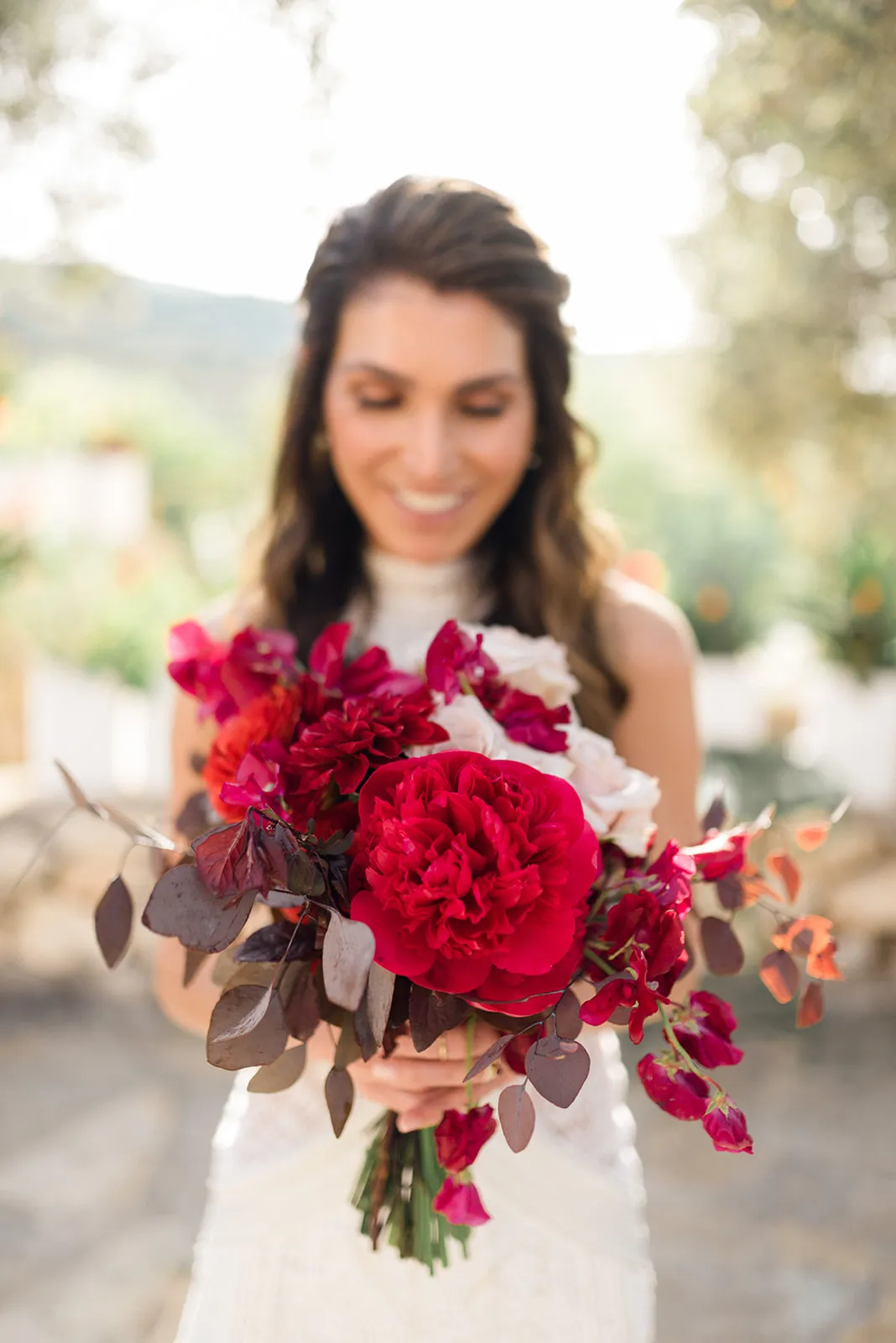 A bride with long wavy hair and a smile, her eyes lowered, holds a lush bridal bouquet forward in soft focus. At its centre sits a large deep red peony, surrounded by ruby-red blooms, creamy white garden roses, and bright orange ranunculus, with trailing dusky eucalyptus leaves. This wedding flower arrangement was created by manfler for a wedding at Agreco Farm in Greece.