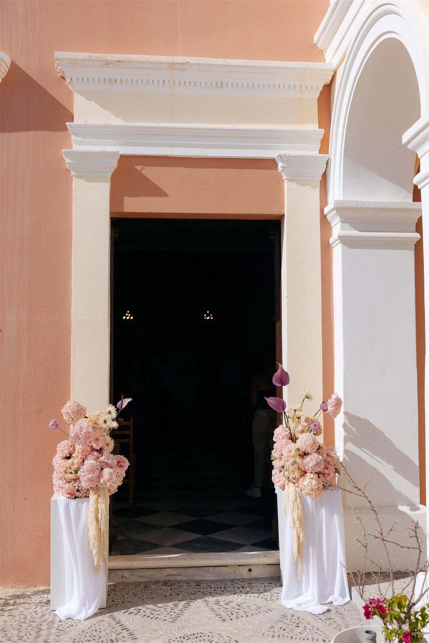 Two tall floral arrangements stand on draped white pedestals either side of a doorway in a peach-coloured wall, framing the dark entrance beyond. Their blooms are soft pink hydrangeas and pale peach flowers, with plum-coloured anthurium, cream pampas-style plumes and small lilac buds. These wedding flowers at Islet in Santorini, Greece, are set against a warm terracotta facade with white cornices and an arched niche, and a black-and-white diamond-tiled floor shows inside the doorway.