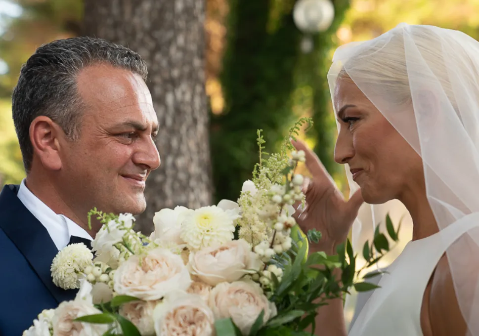 A bridal bouquet of soft blush garden roses, creamy white dahlias and sprigs of white astilbe and berries, with green foliage trailing from it, held close as a bride in a sheer veil gazes at her smiling groom in a navy suit. Behind them is a blurred green garden with a tree trunk. This is a wedding flower moment at SAPEL HALL in Agia Marina, Crete, Greece.