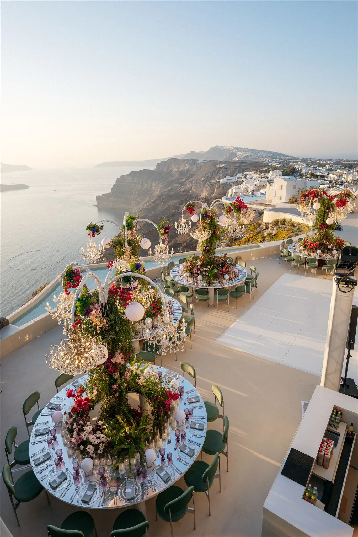 Round tables on a clifftop terrace at Islet Santorini in Thira, Greece, are dressed for a wedding dinner, each with a tall centrepiece of red bougainvillea, garden roses, pink and white blooms and trailing green foliage beneath arched crystal chandeliers and white globe lanterns. Green velvet chairs surround the tables, and beyond the terrace the caldera drops to a calm sea under a pale, sunset-tinted sky, with whitewashed village houses clinging to the cliff in the distance.