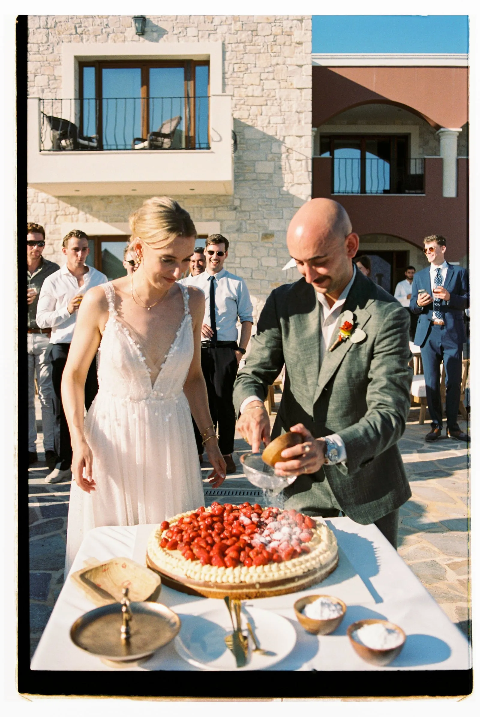 A bride in a sleeveless, V-neck lace gown and a groom in a grey-green suit with a small floral boutonniere stand at a white-draped table on a stone terrace, where he is grating sugar over a round cake topped with fresh strawberries. Guests in shirts and blazers watch from behind them under warm late-afternoon light. This is a wedding in Crete, Greece, at Wine Dark Sea Villas in Rodia.