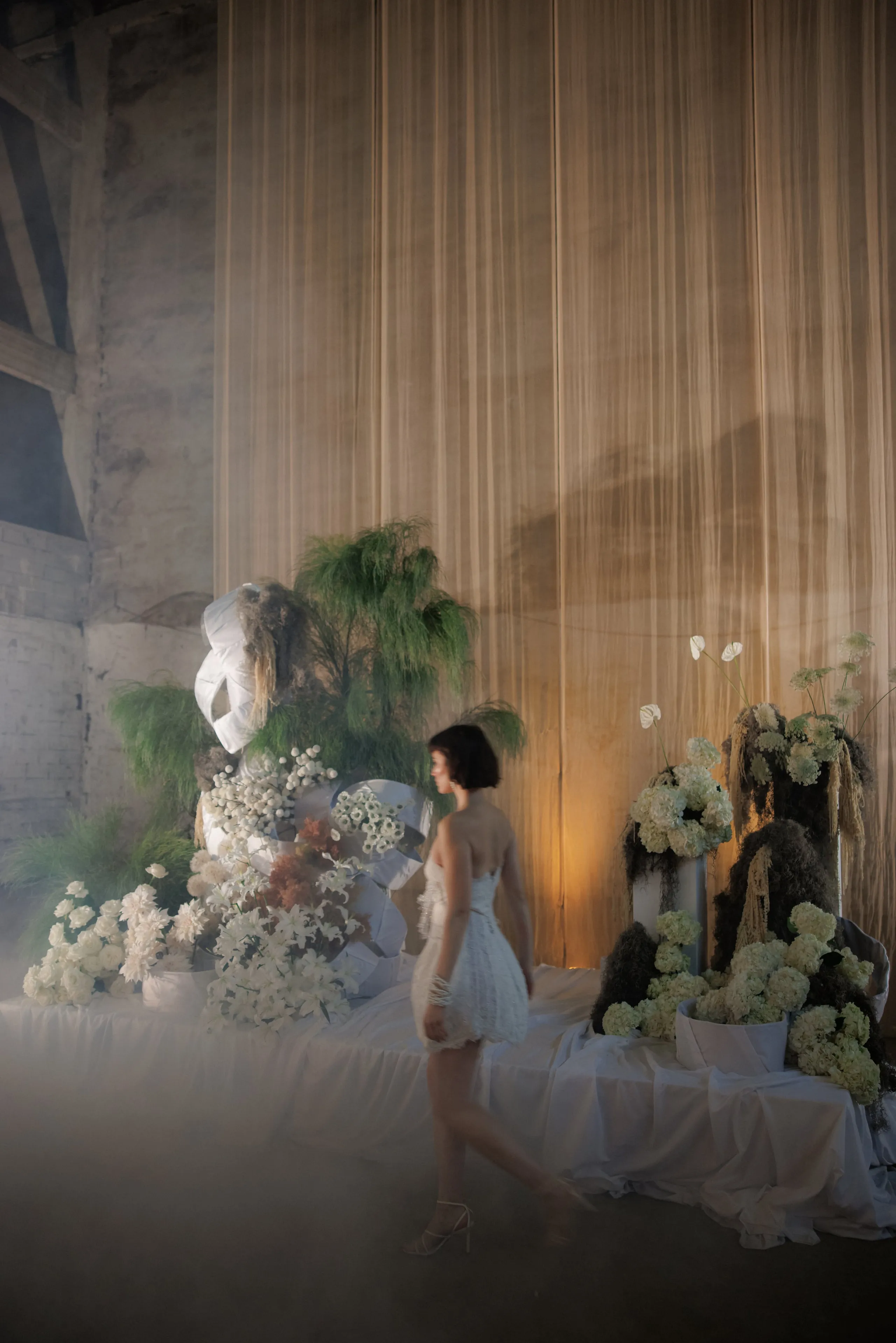A woman in a short, pale strapless dress with fringed detailing walks past a low white draped platform in a dim, hazy hall at Rethimno, Crete, Greece. Behind her, tall cascades of white flowers, including what look like white orchids, hydrangeas and white anthuriums, spill over large pots and sculptural white forms, with feathery green fern-like foliage rising above. Soft amber light falls through a sheer gold curtain, making the scene feel dreamy and ethereal, in this event floral installation.